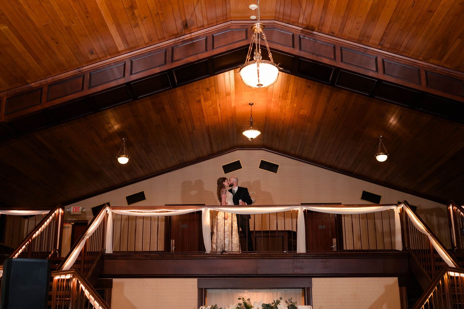 Bride and groom kissing on indoor balcony of rustic wood-ceiling wedding reception venue.