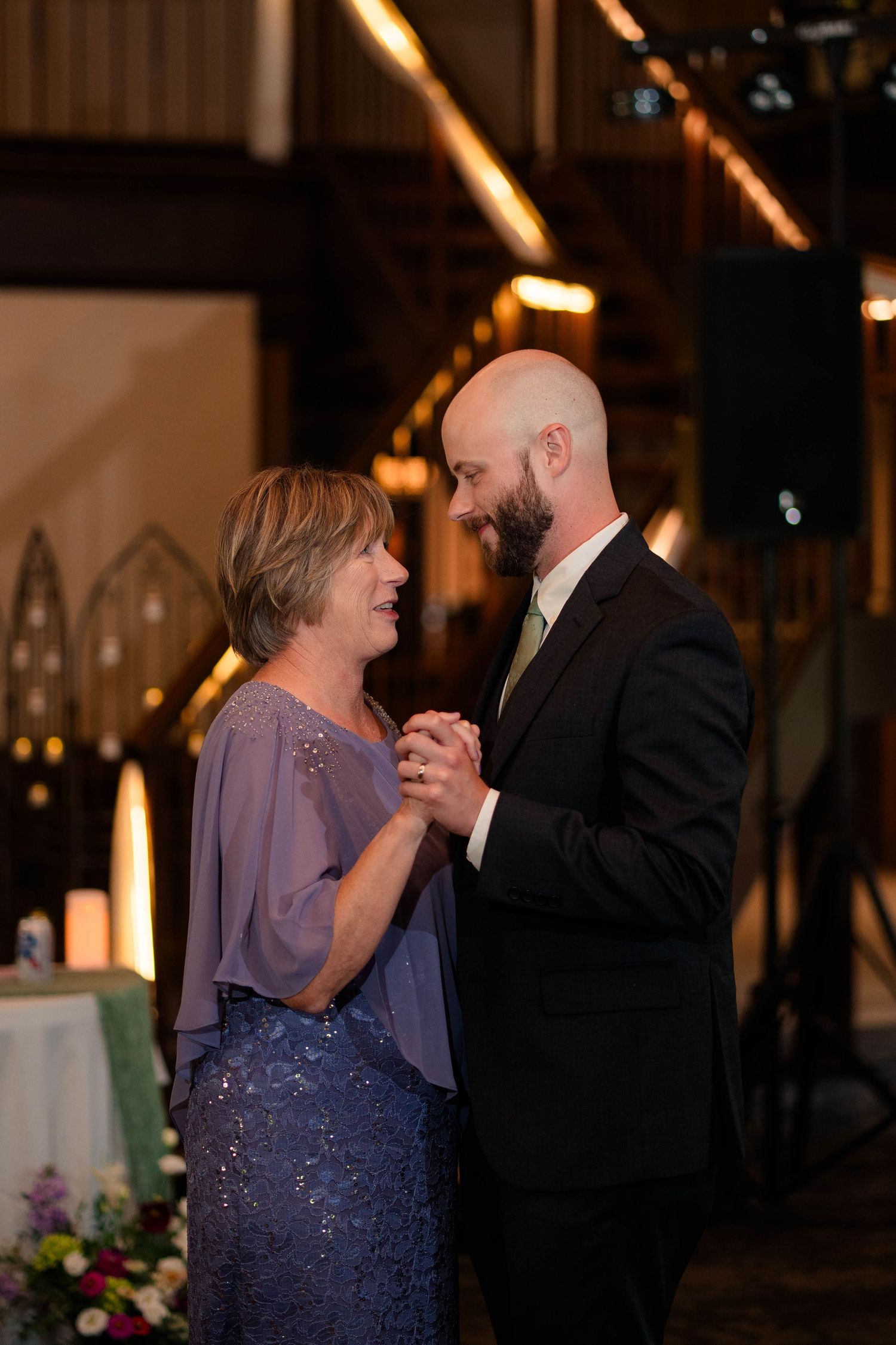 Bald groom with beard shares emotional mother-son dance at wedding reception in warmly lit venue.