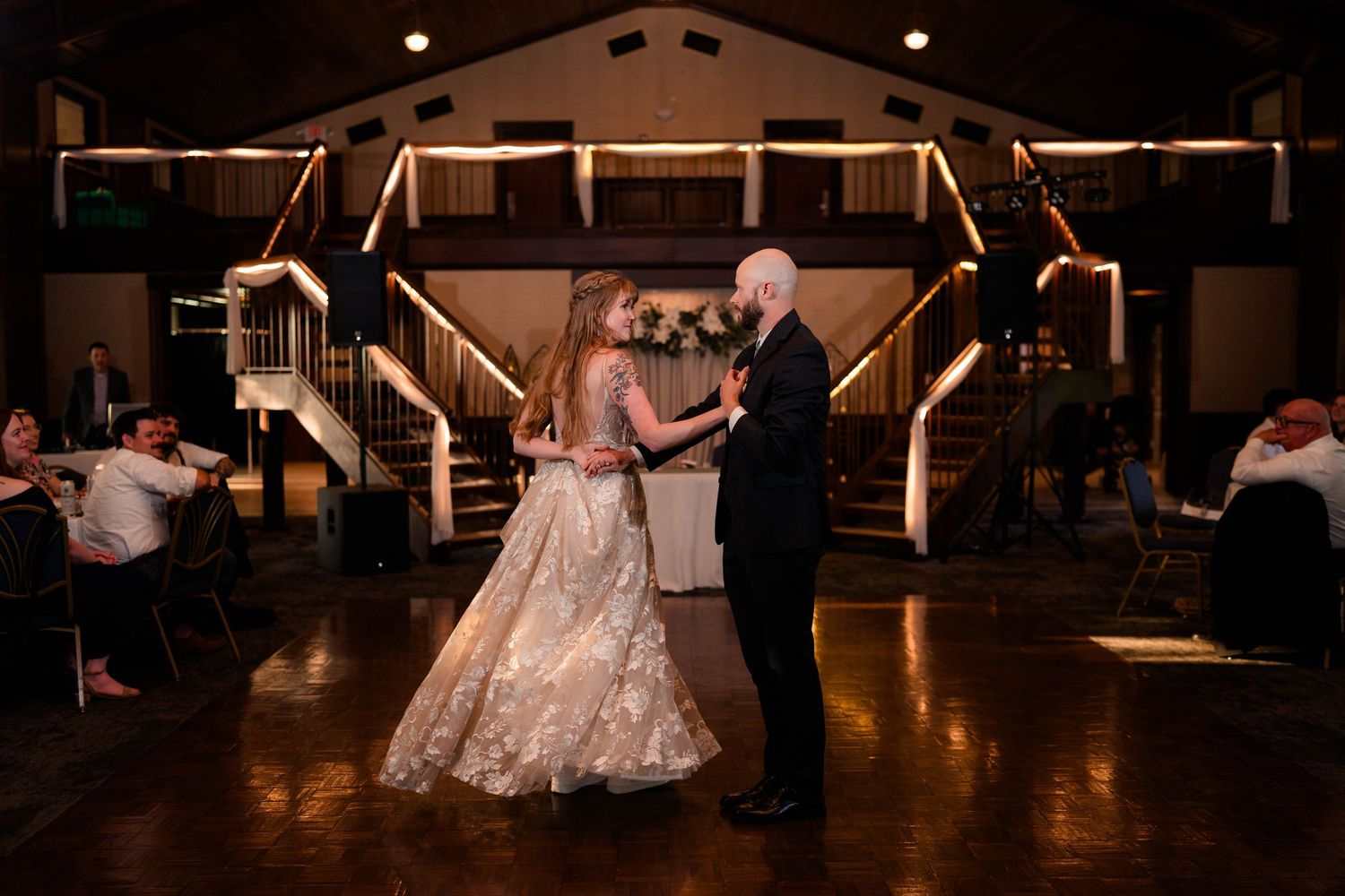 Bride in flowing lace gown twirls with partner during elegant first dance at indoor wedding reception.