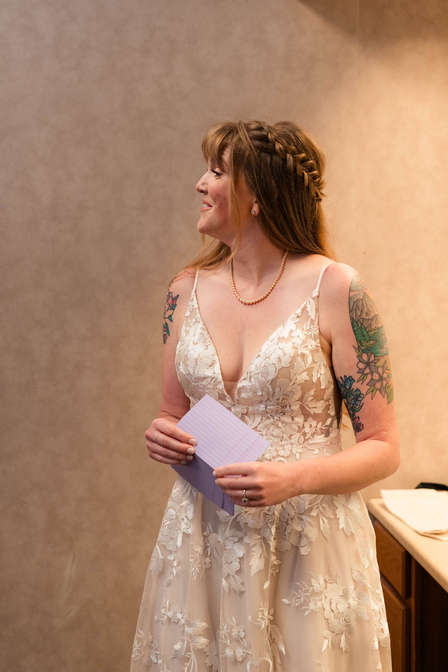 Smiling bride in lace wedding gown with tattoos holding vows card indoors.
