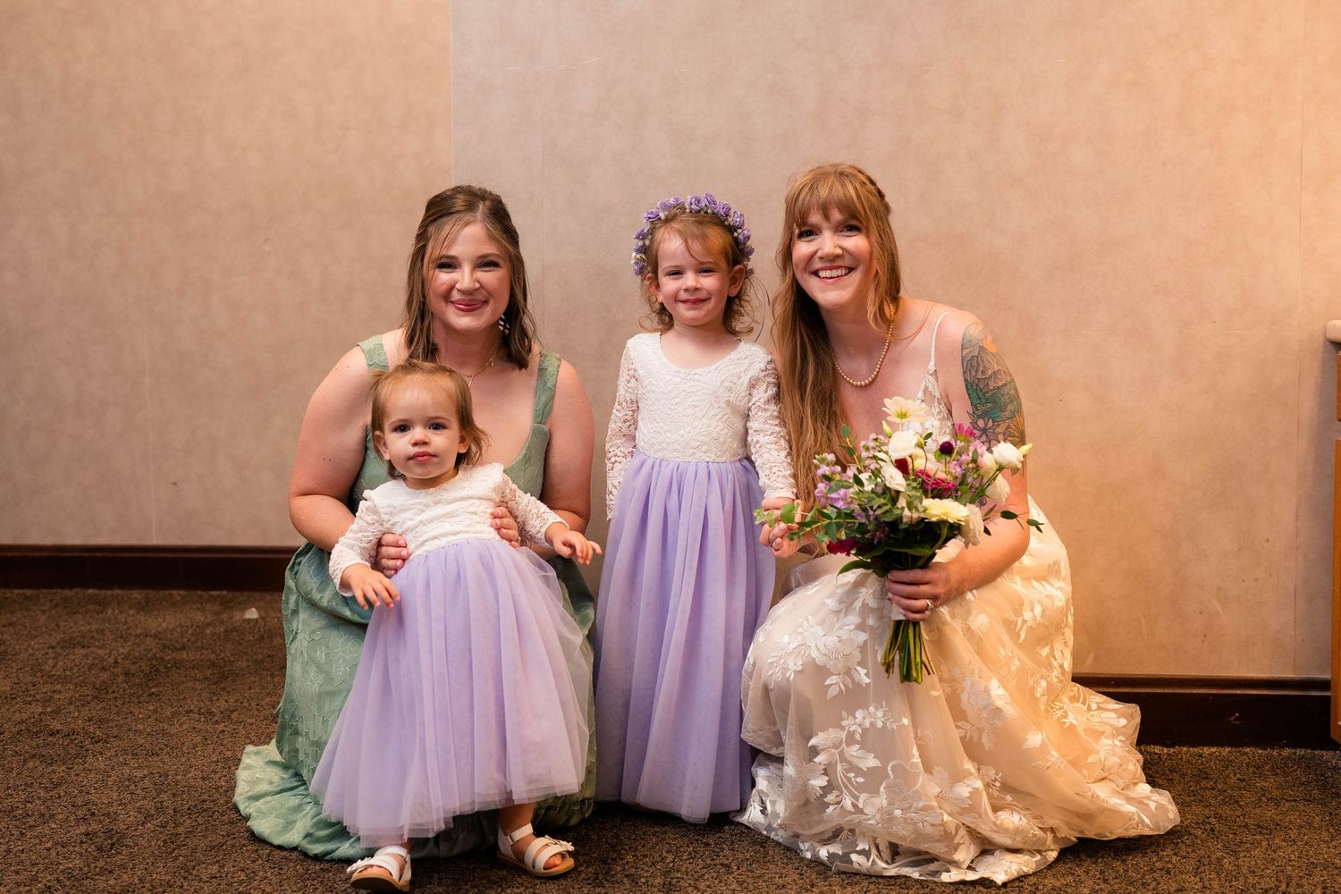 Bride in lace gown kneels with two flower girls in purple dresses and woman in sage green bridesmaid dress.
