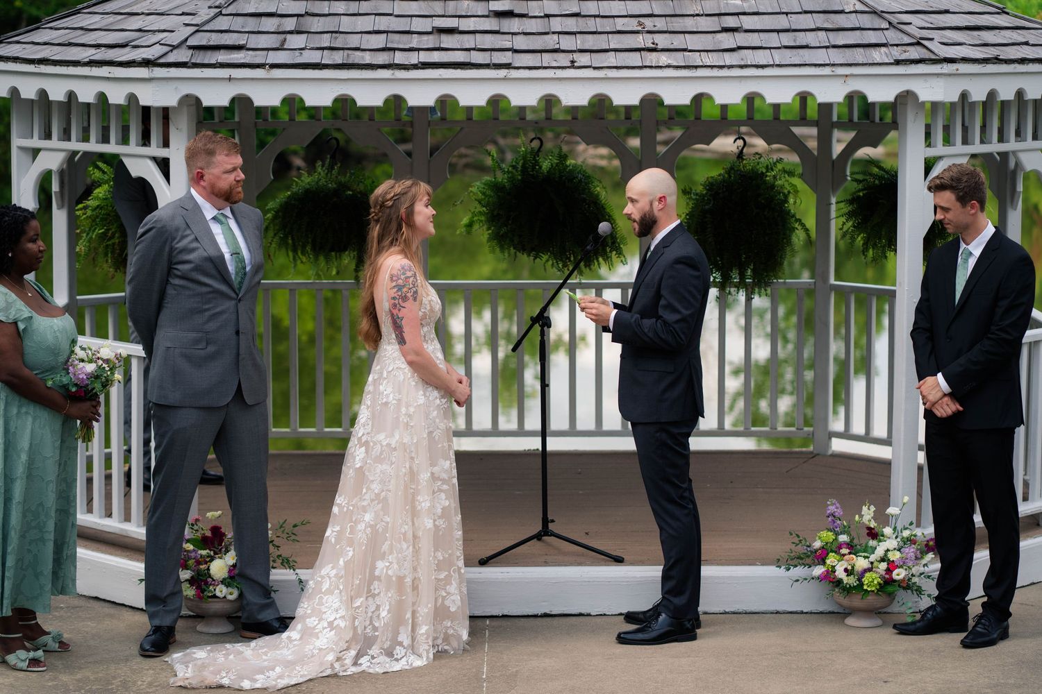 Bride and groom exchange vows at white gazebo altar with officiant and wedding party during outdoor ceremony.