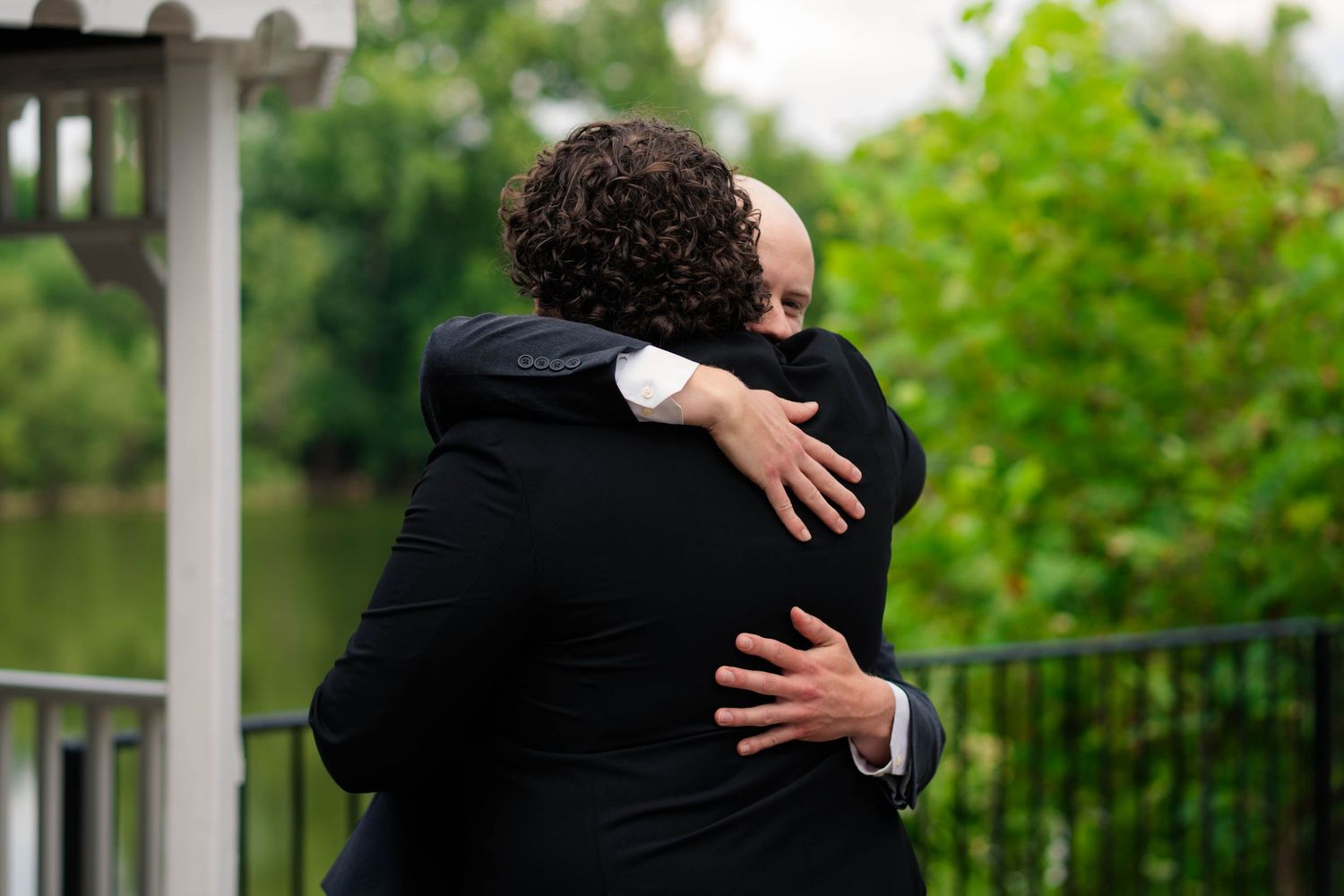 Two wedding guests share a warm embrace on an outdoor gazebo near a lake.