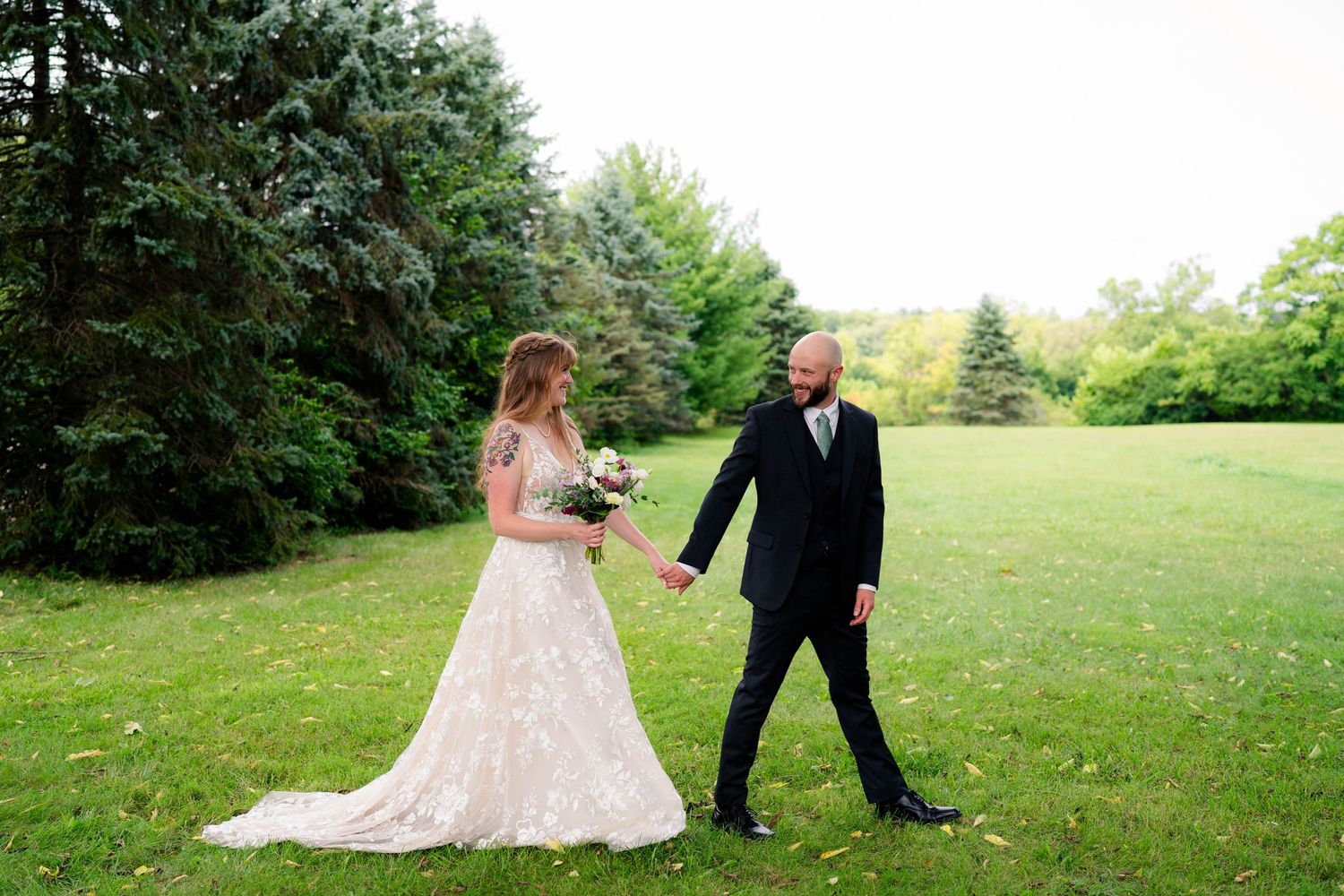 Bride in lace gown and groom in black suit walk hand in hand across green lawn at outdoor wedding.