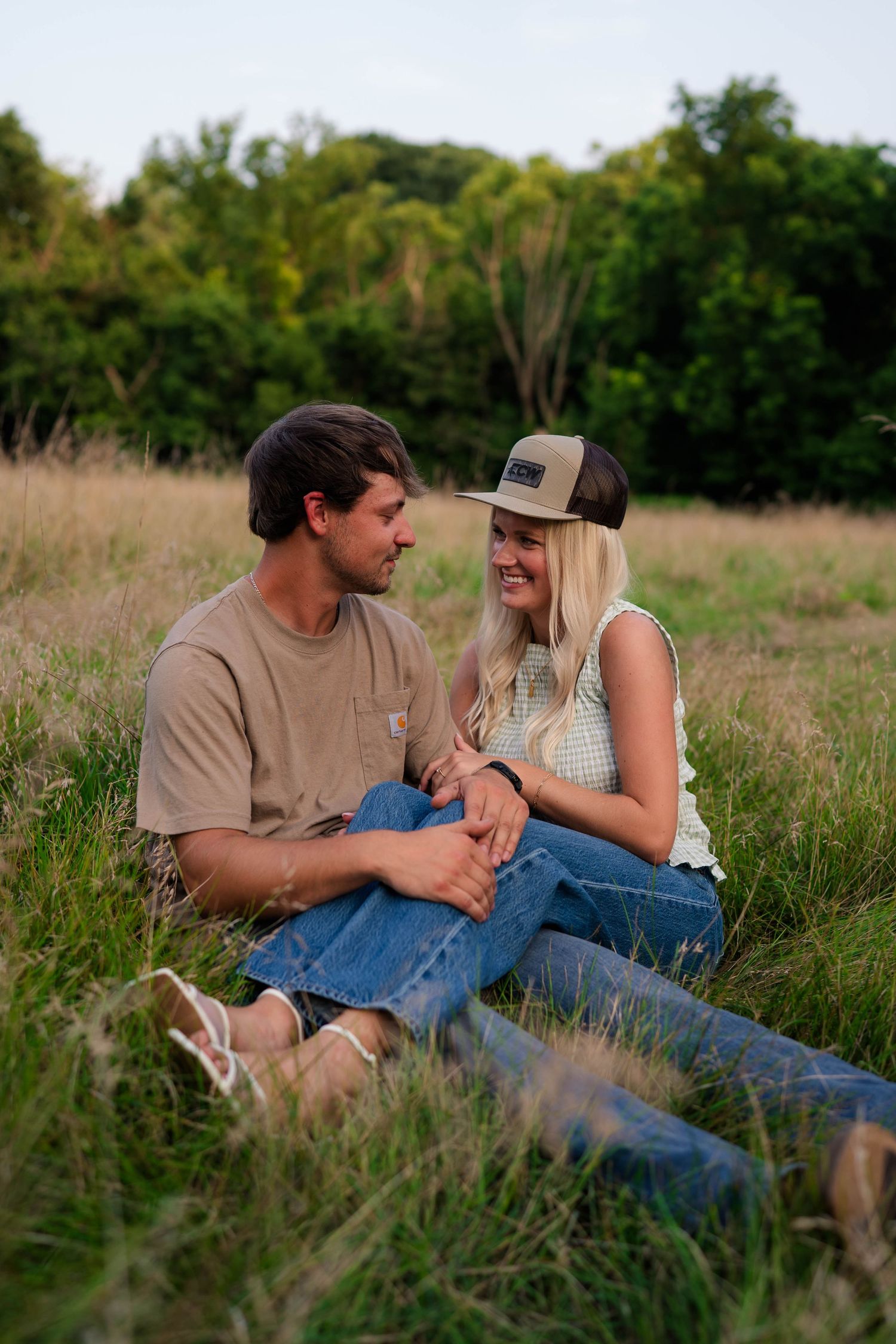 A couple sits together in a grassy field, smiling at each other, woman wearing a cap and casual outfit.