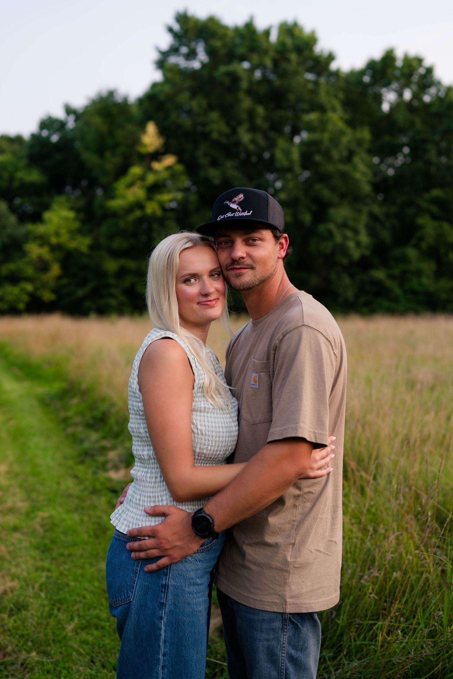 A couple embraces on a grassy path, woman in checkered top and jeans, man in beige shirt and black cap.