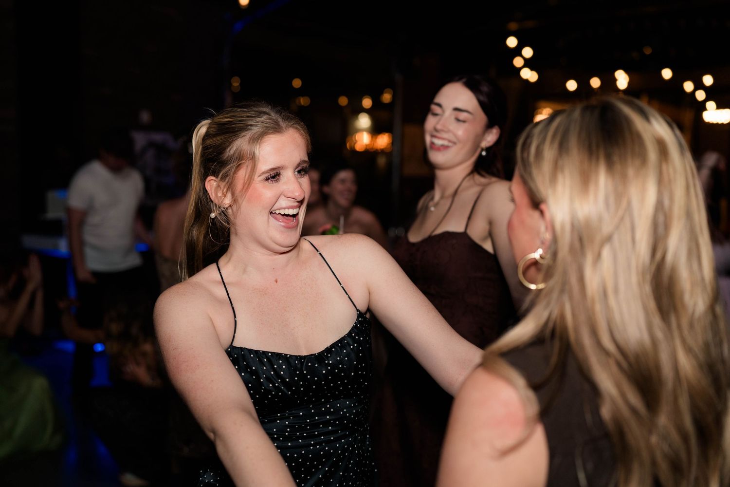 Women laughing and dancing together at an evening wedding reception under string lights.