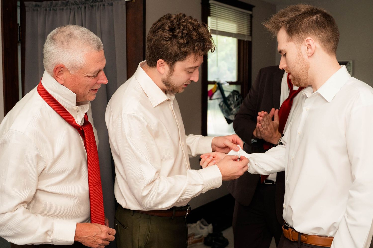 Three men in white dress shirts and red ties helping each other prepare, likely groomsmen getting ready for a wedding.