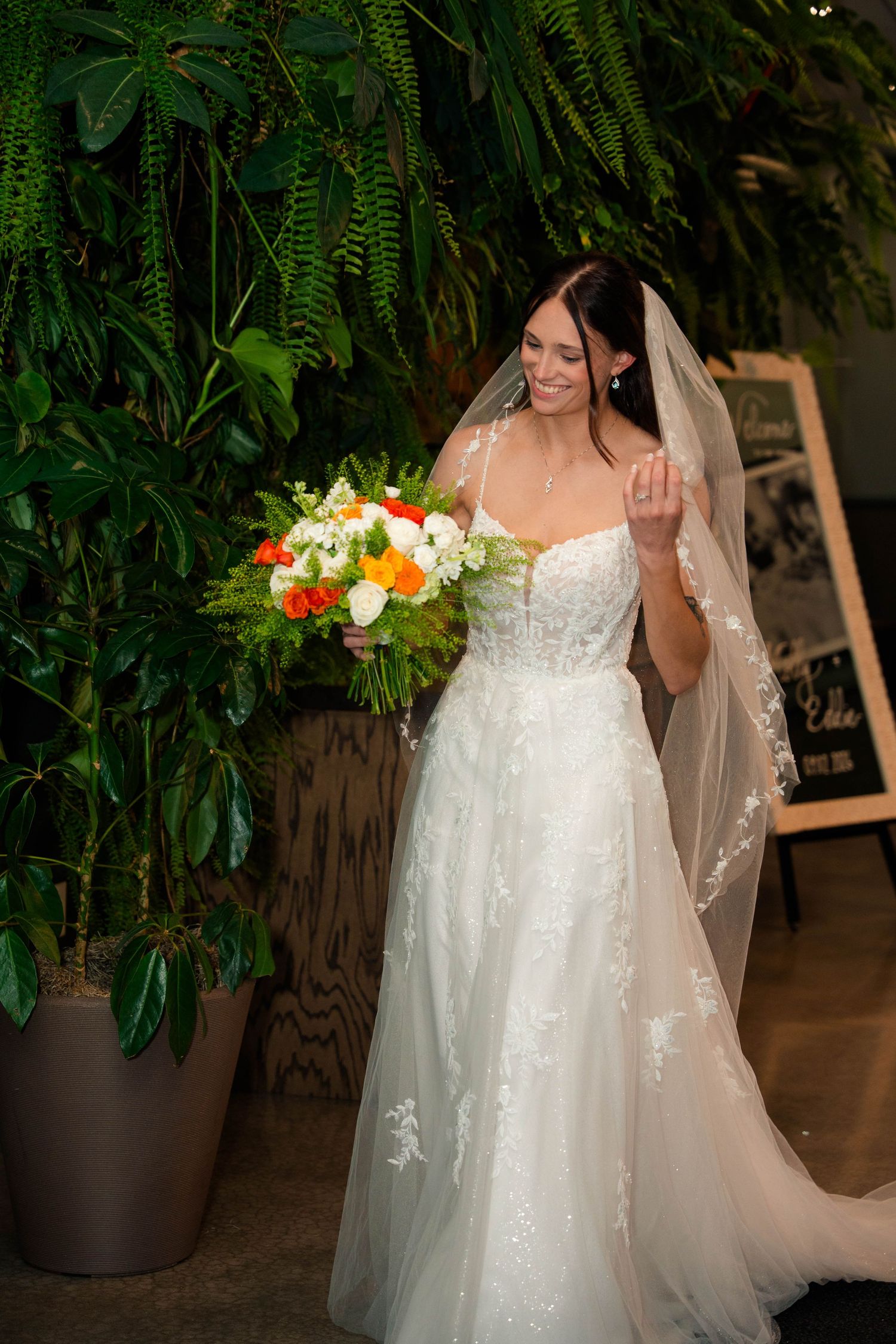 Bride in lace A-line gown holding colorful bouquet standing among lush greenery.