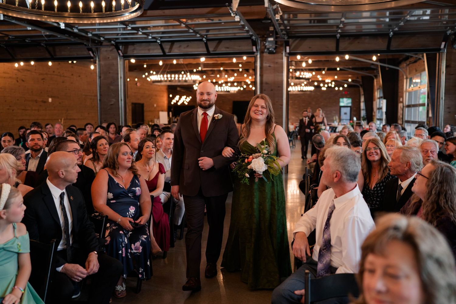 Bridesmaid in emerald green dress walks down aisle with escort at crowded wedding ceremony.