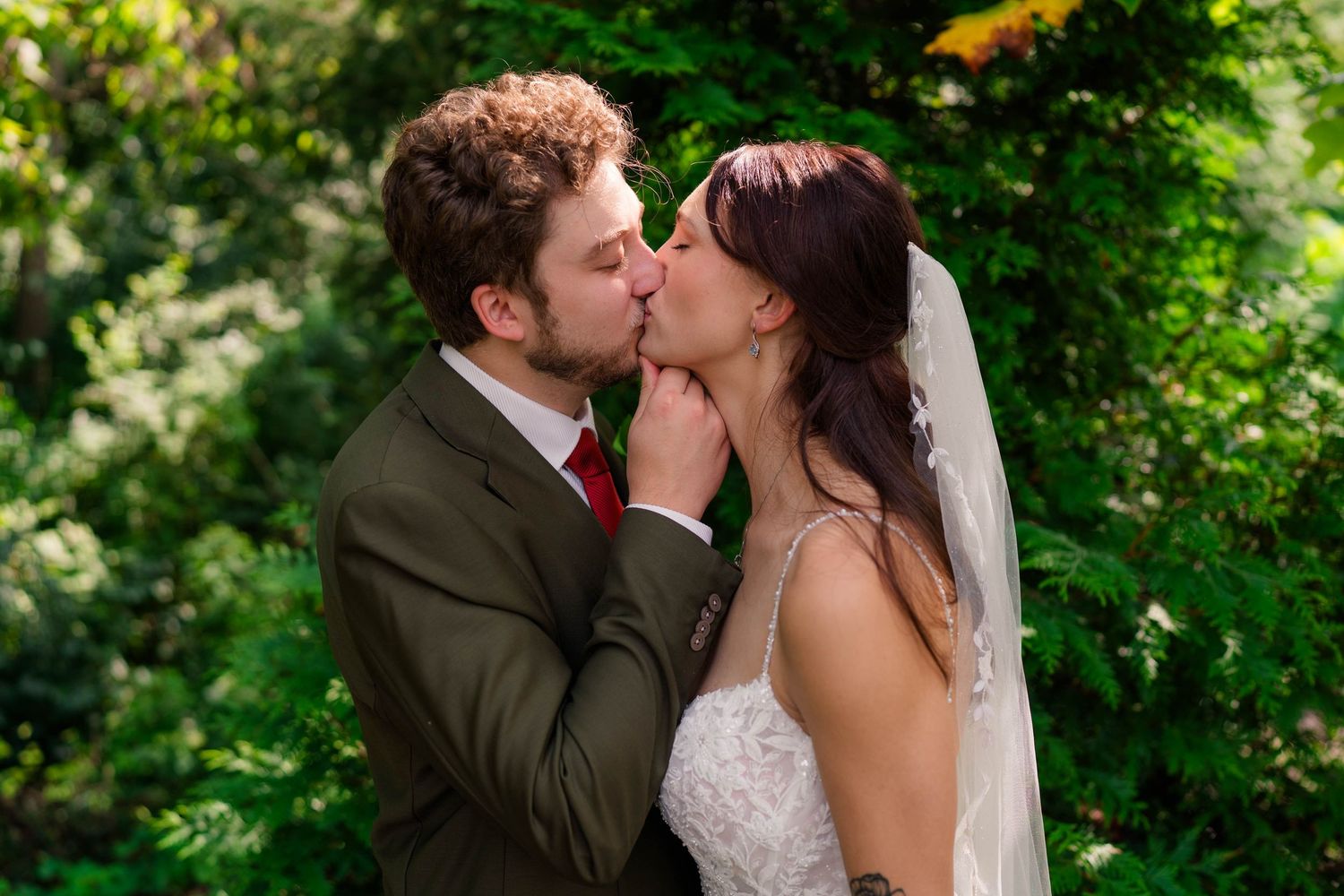 Bride and groom sharing a romantic kiss outdoors surrounded by lush green foliage on their wedding day.