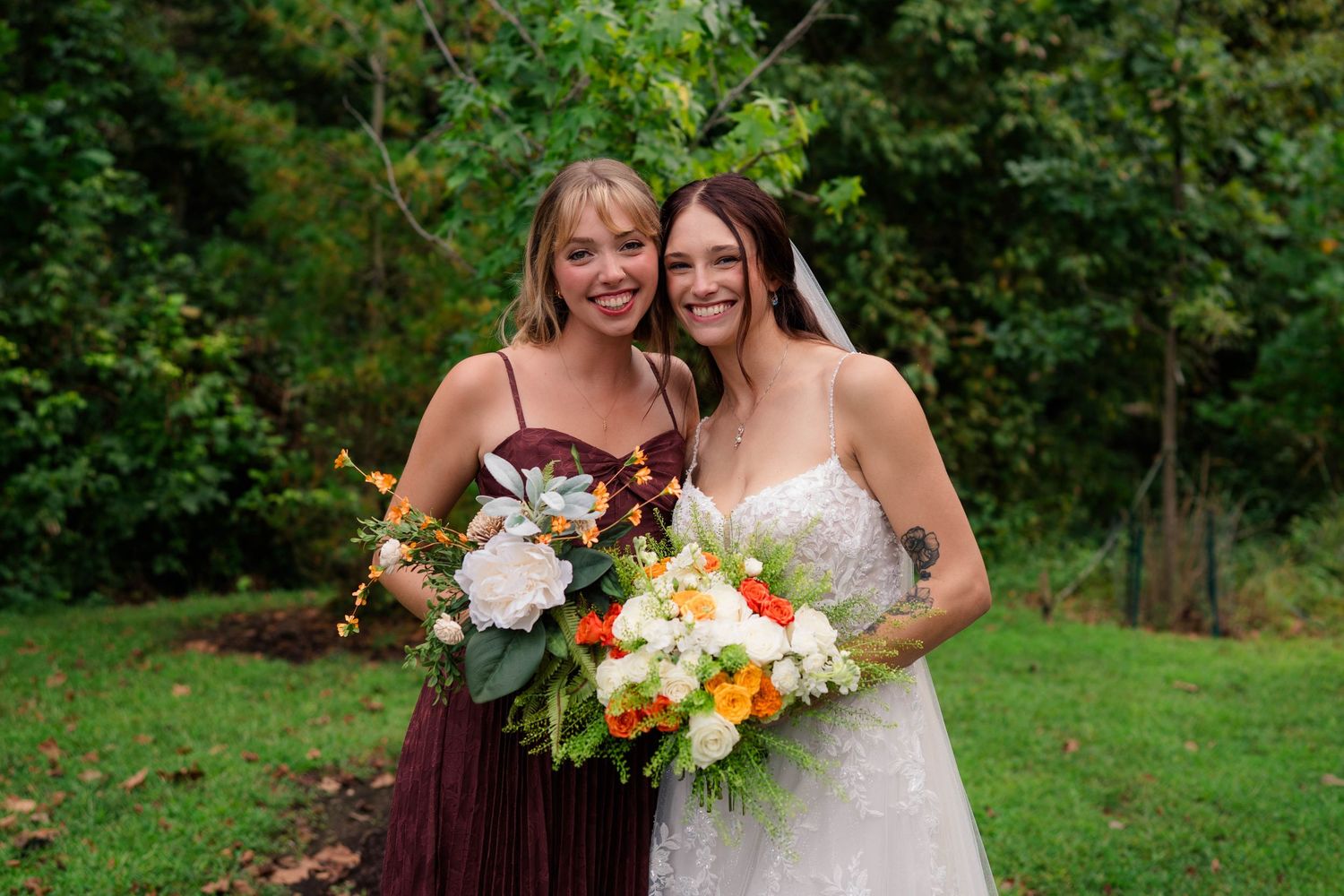 Bride and bridesmaid in burgundy dress smile together outdoors holding beautiful white and orange bouquets.