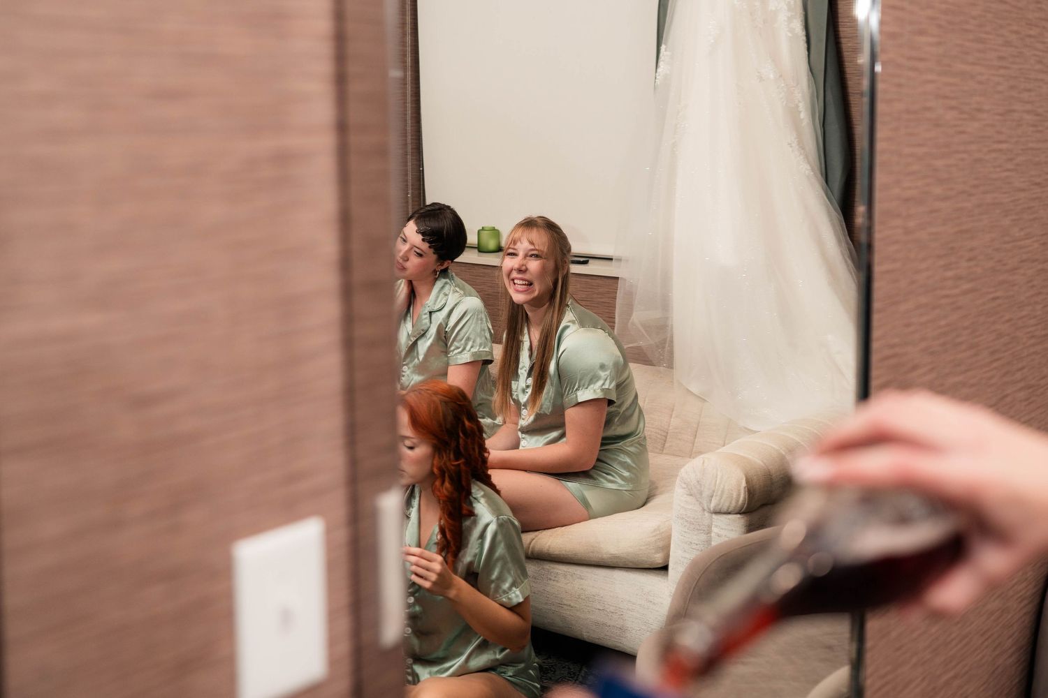 Bridesmaids in sage robes laugh together while getting ready for a wedding.