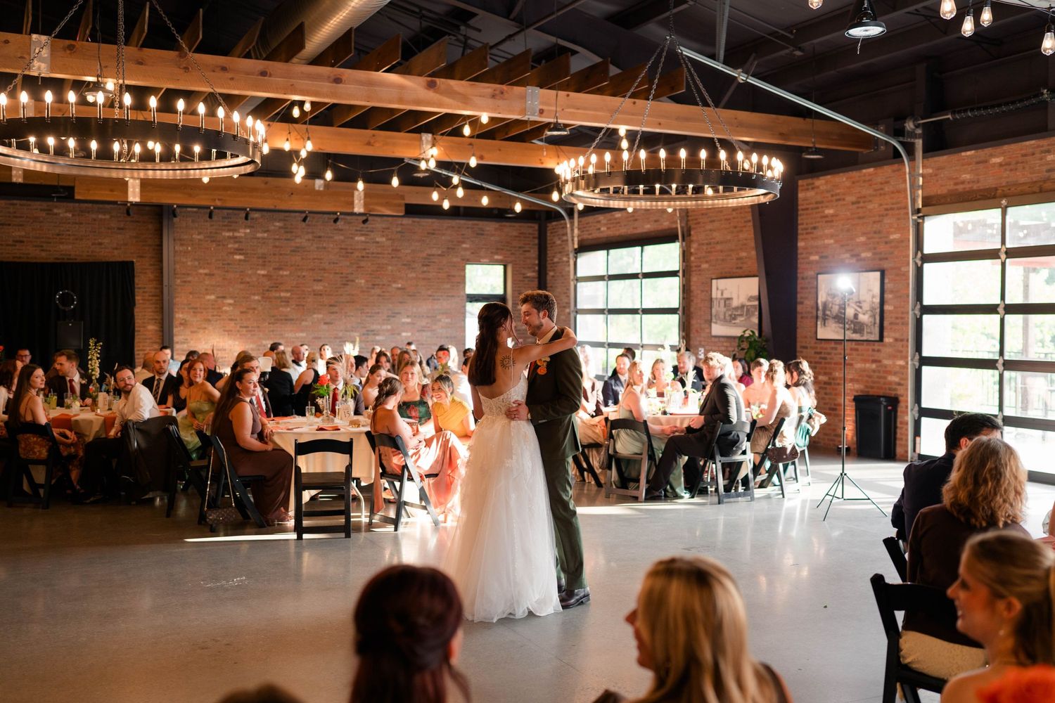 Bride and groom share first dance in rustic industrial wedding venue with chandelier lighting.