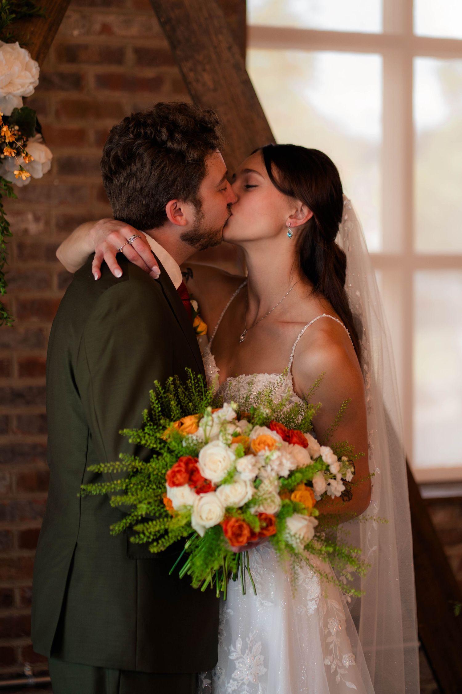 Newlyweds share first kiss at altar in rustic brick wedding venue with wooden arch and floral decor.