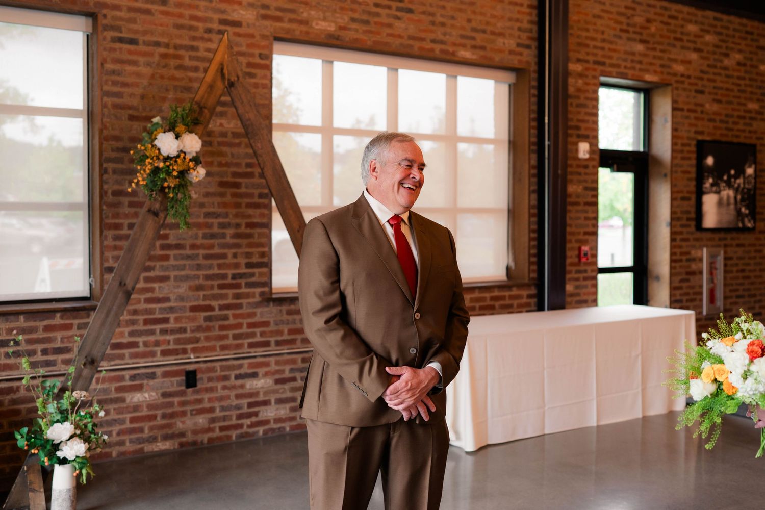 Father of the bride smiling proudly at rustic brick wedding venue with floral arch.