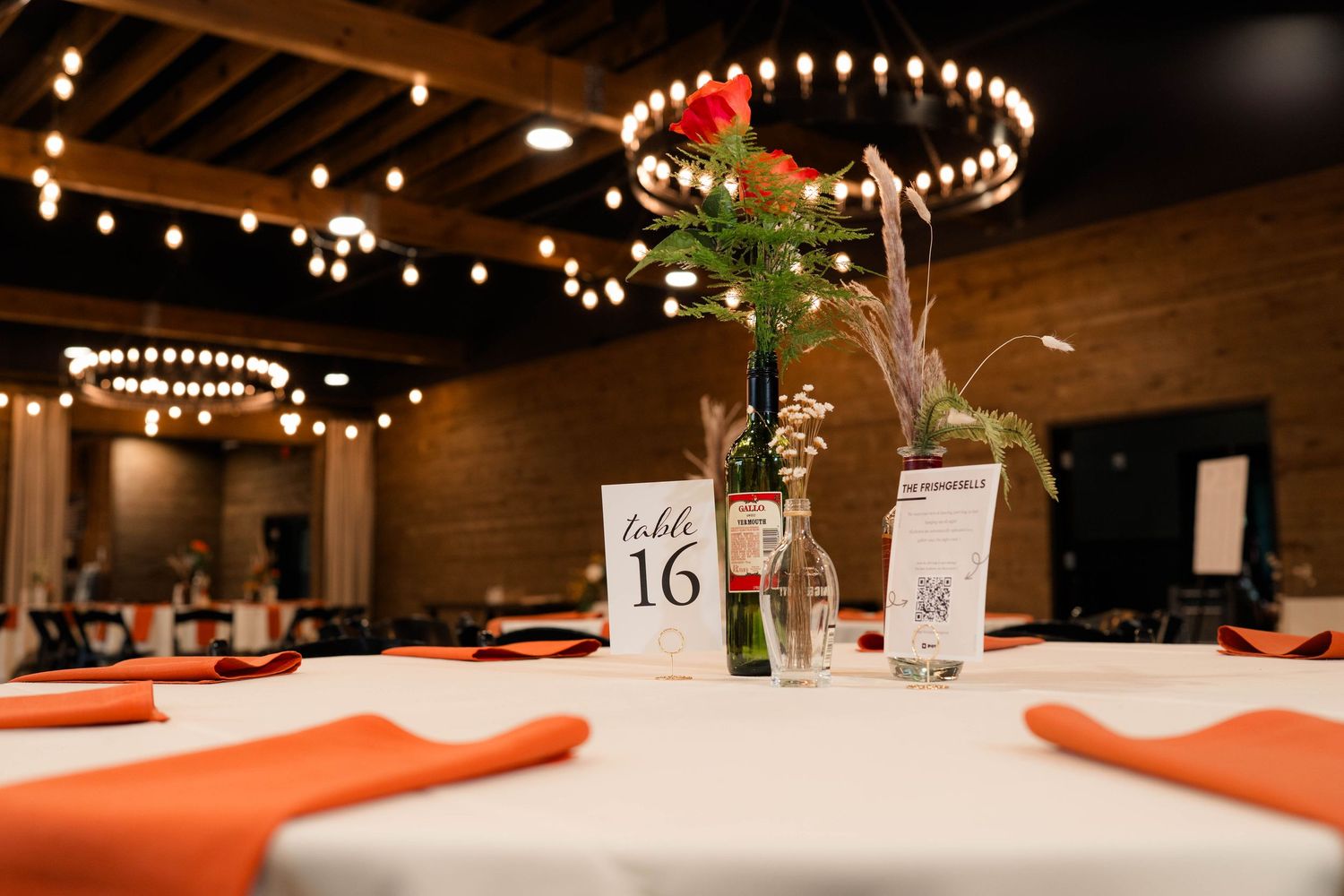 Wedding reception table 16 with orange napkins, floral centerpiece, and string lights overhead.