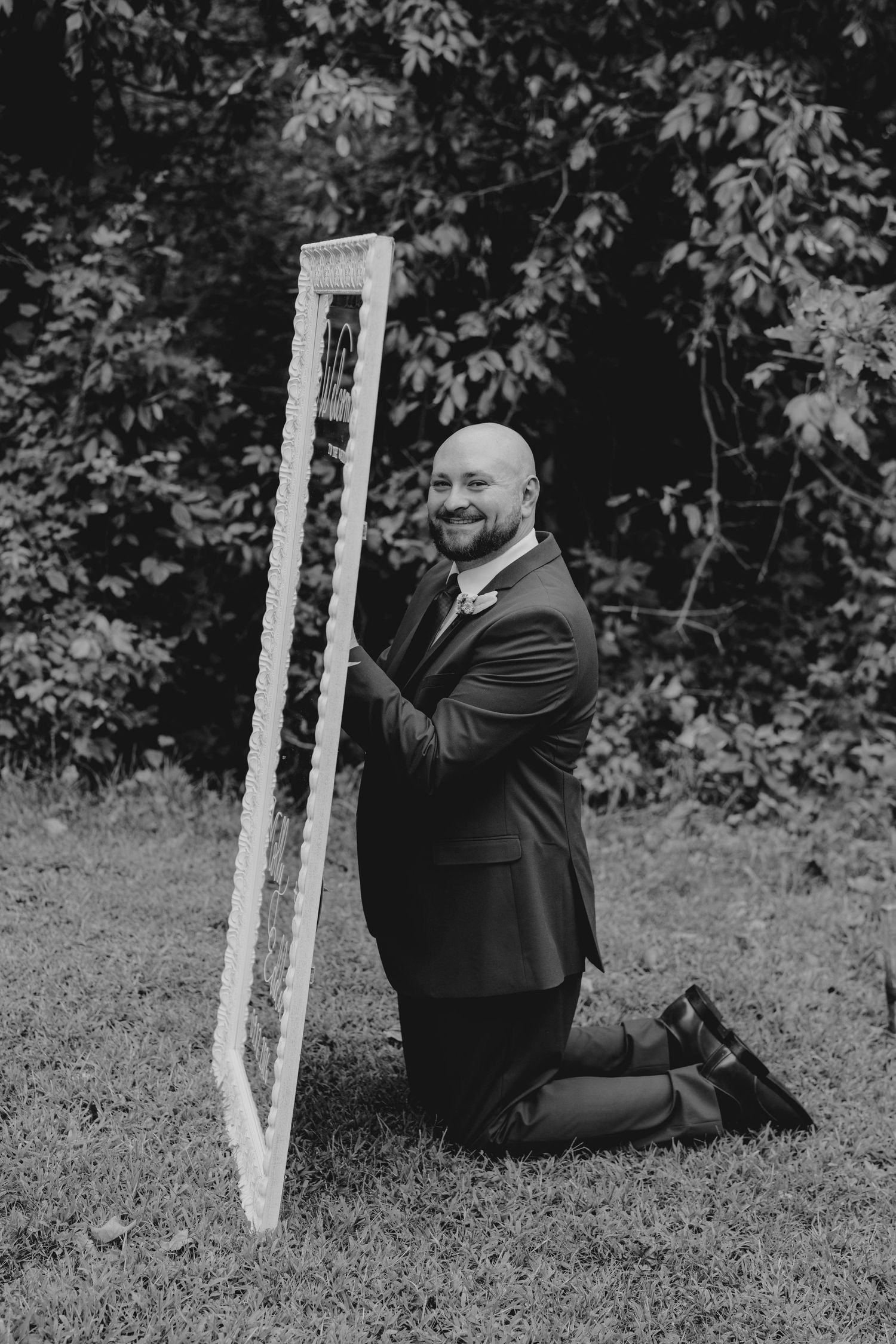 Groom in dark suit kneeling on grass holding a decorative frame in a black and white wedding photo.