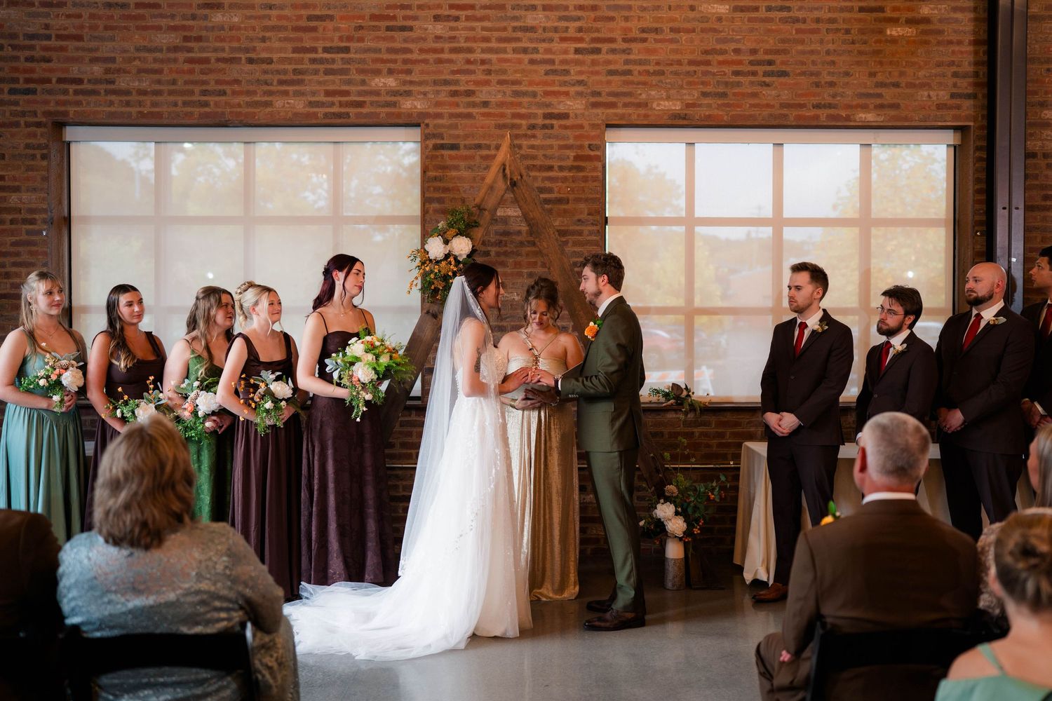 Bride and groom exchange vows at altar surrounded by wedding party in brick-walled indoor venue.