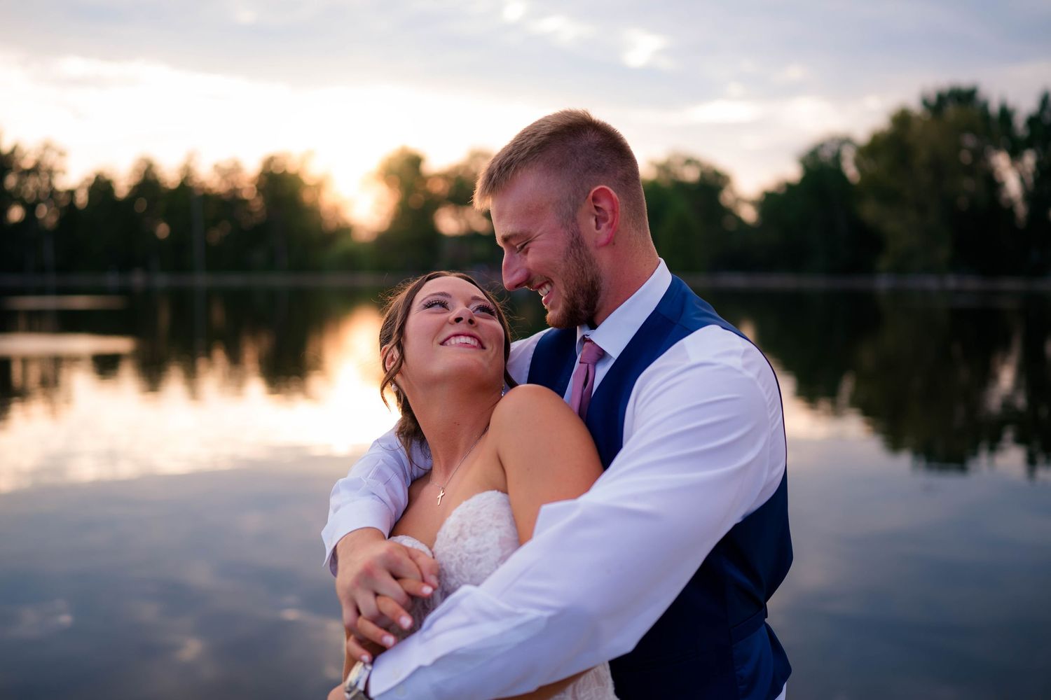 Bride and groom embracing by a lake at sunset, sharing a romantic moment with golden light reflecting on the water.