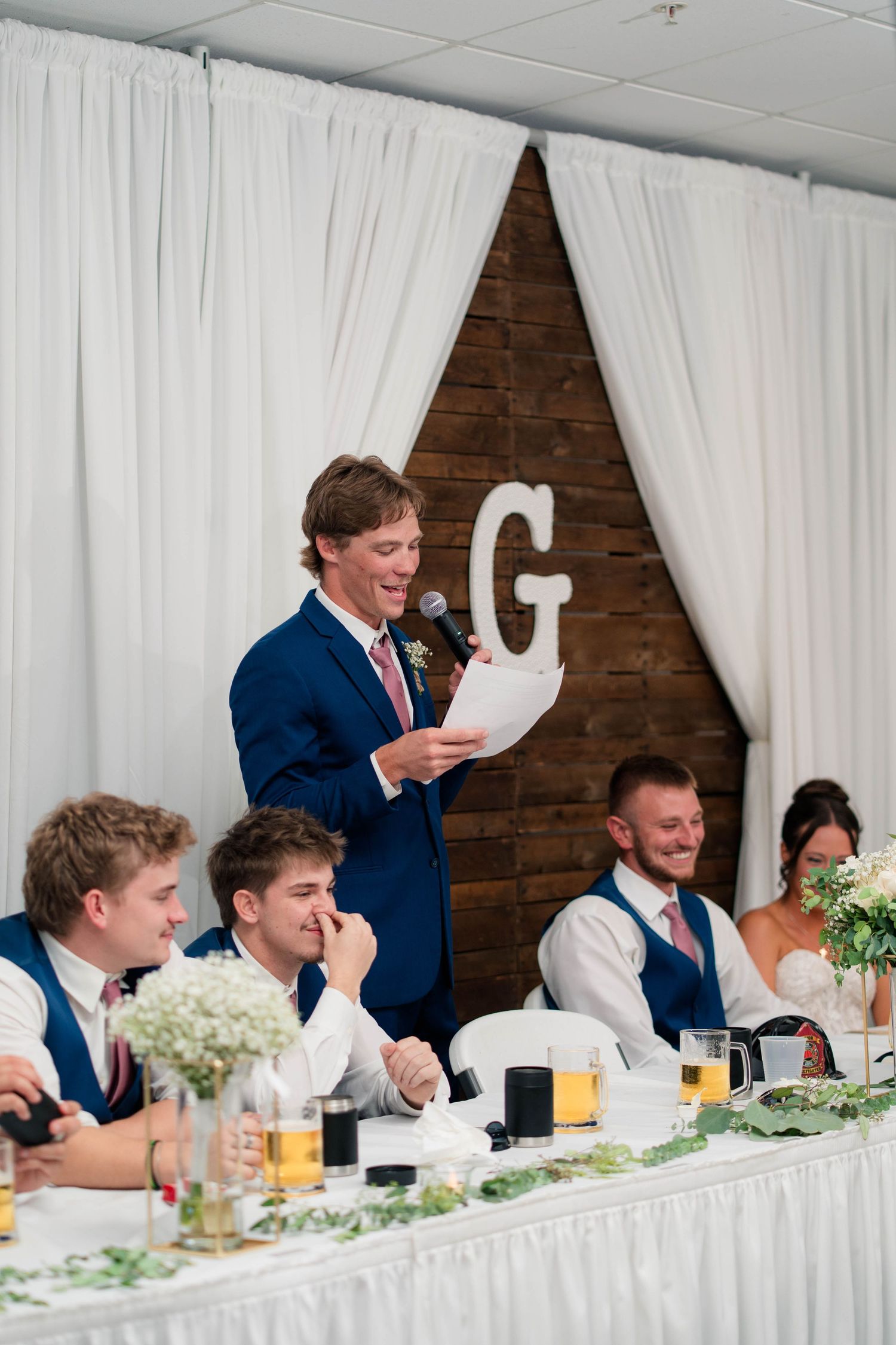 Best man delivers speech at wedding reception while groom and bridal party laugh at the head table.