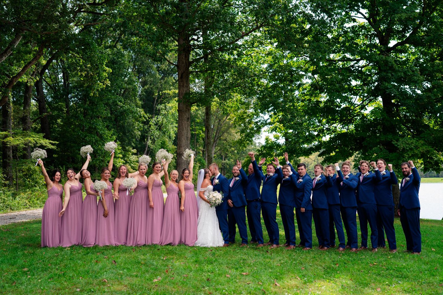 Large wedding party poses outdoors, bridesmaids in mauve gowns and groomsmen in navy suits celebrating among lush green trees.