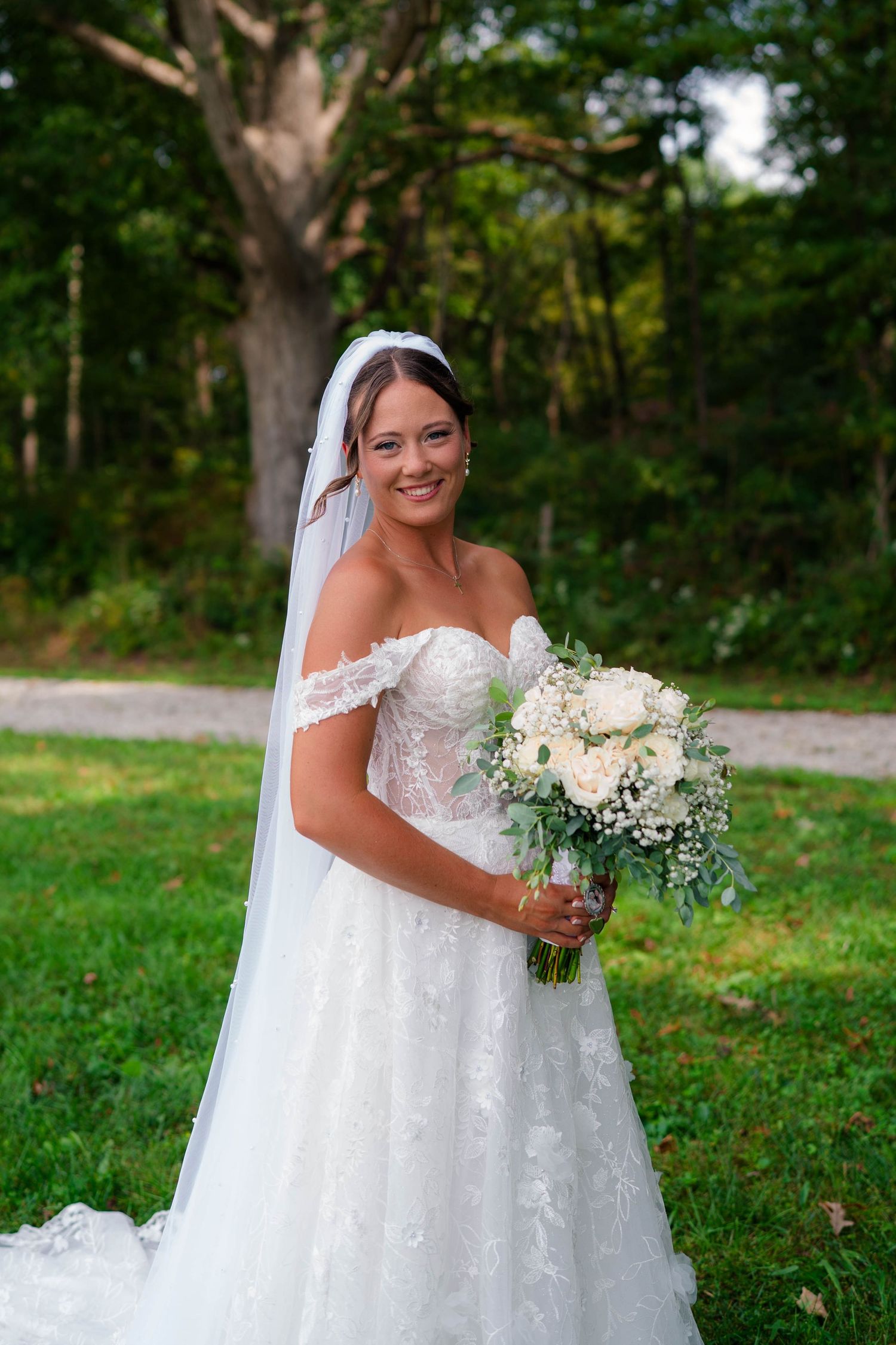 Beautiful bride in off-shoulder lace wedding gown smiles holding a white rose bouquet outdoors.