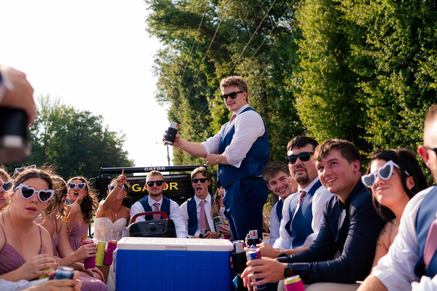 Groomsman stands on decorated wagon holding drinks while guests in sunglasses celebrate at outdoor wedding reception.