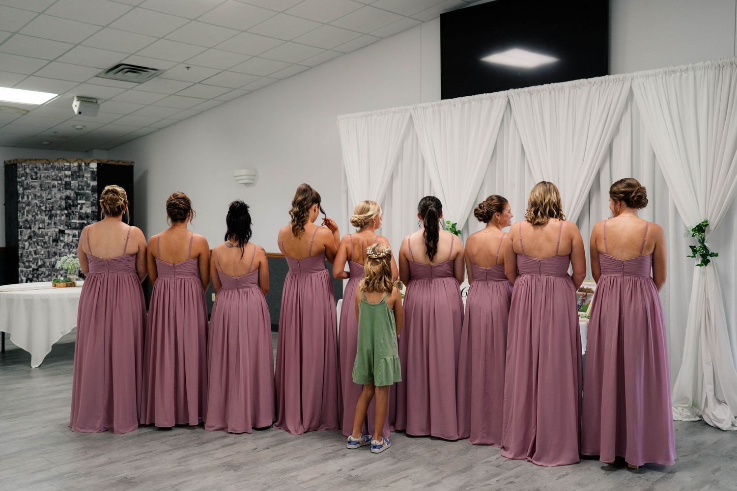 Eight bridesmaids in mauve gowns stand with backs to camera beside a small child in green dress at a wedding venue.