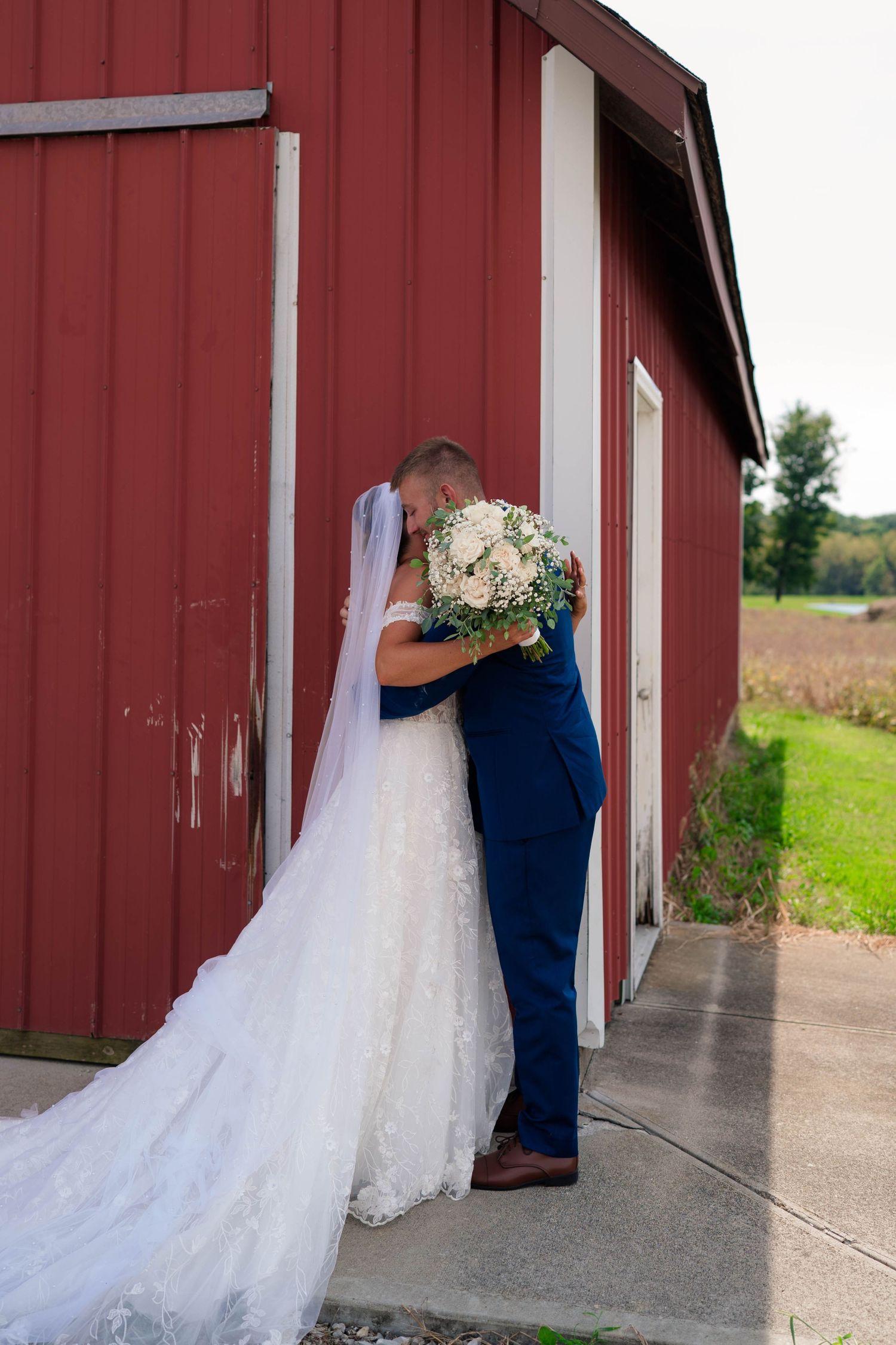Bride and groom sharing romantic kiss outside red barn on wedding day with white bouquet.