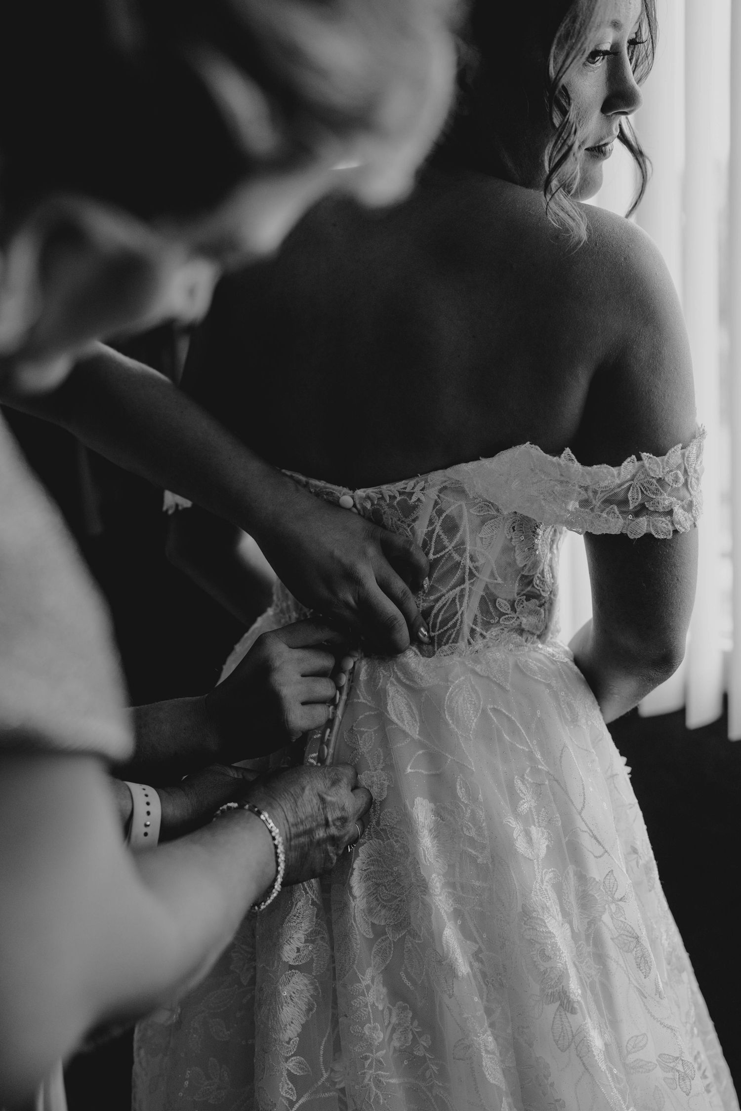 Assistant helping bride fasten buttons on off-shoulder lace wedding gown in black and white photo.