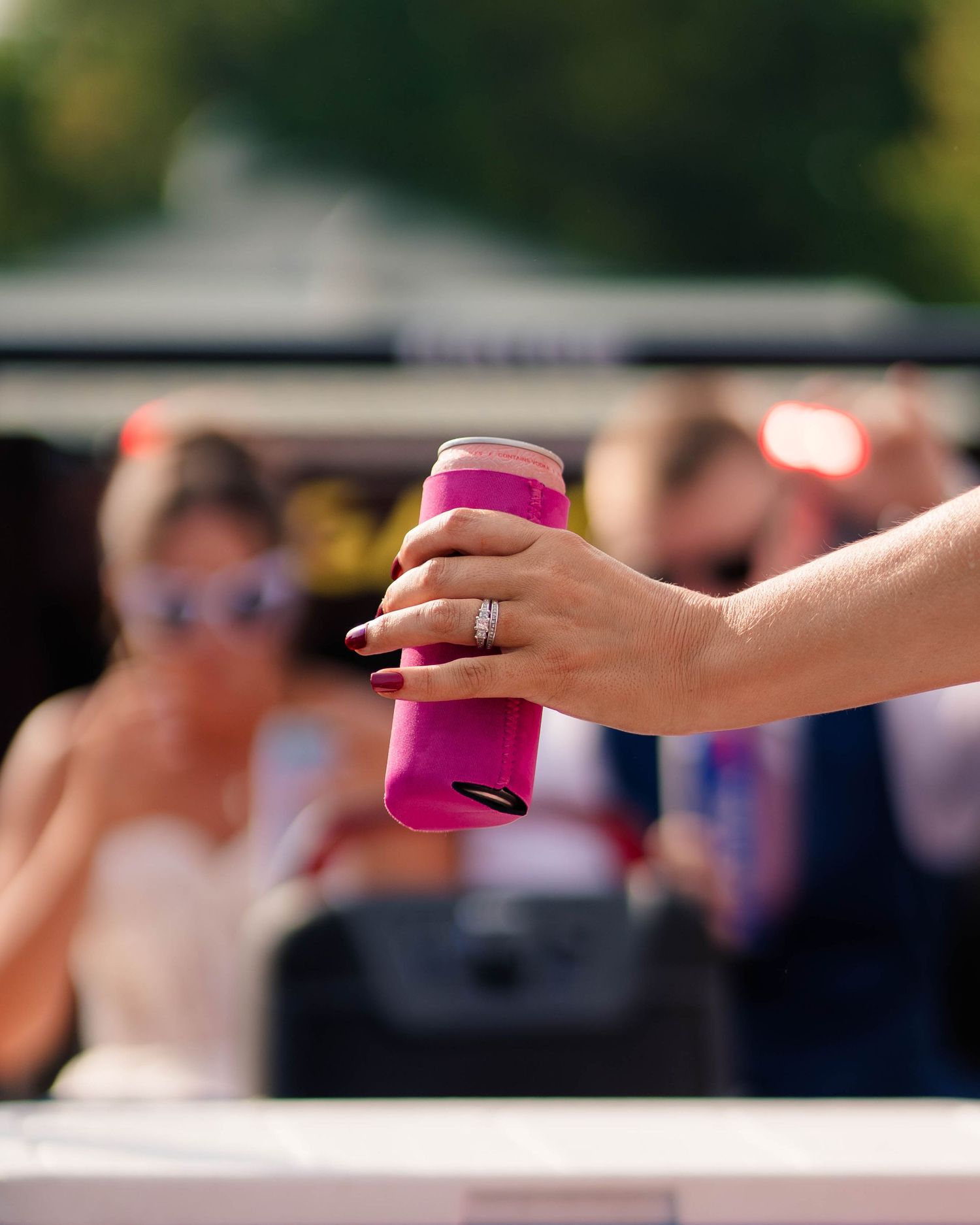 Close-up of a hand with an engagement ring holding a pink koozie at an outdoor wedding reception.