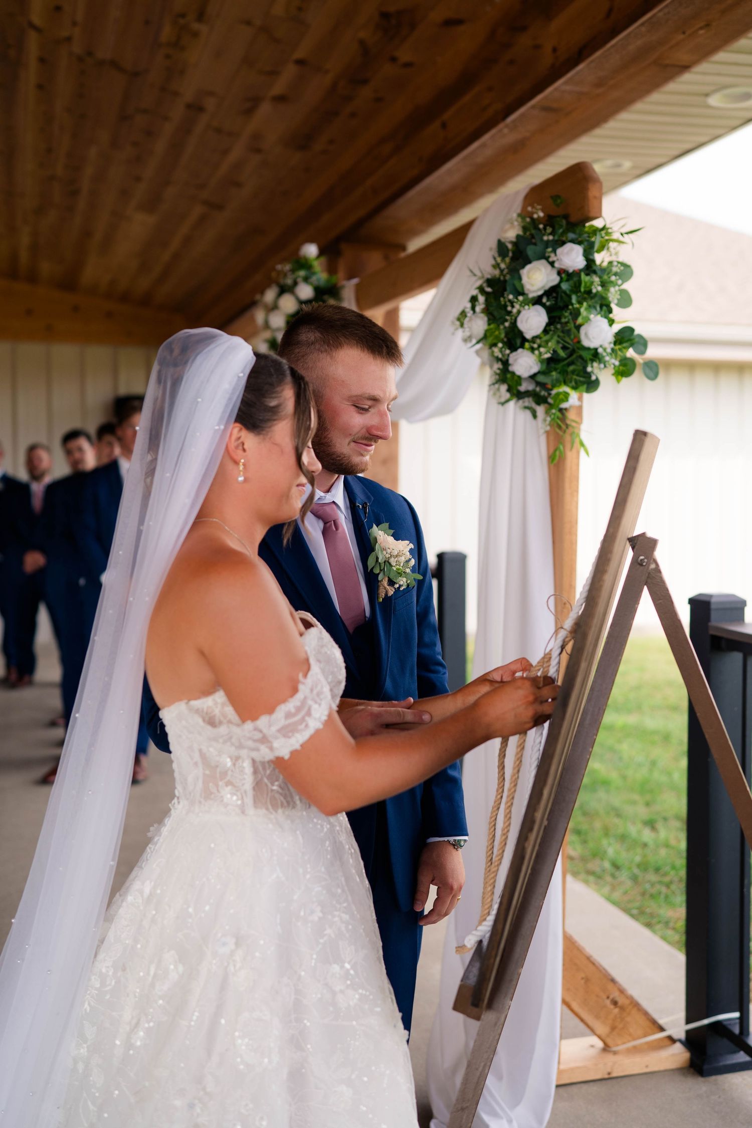 Bride and groom participate in unity painting ceremony at outdoor wedding under wooden pavilion with floral arch.