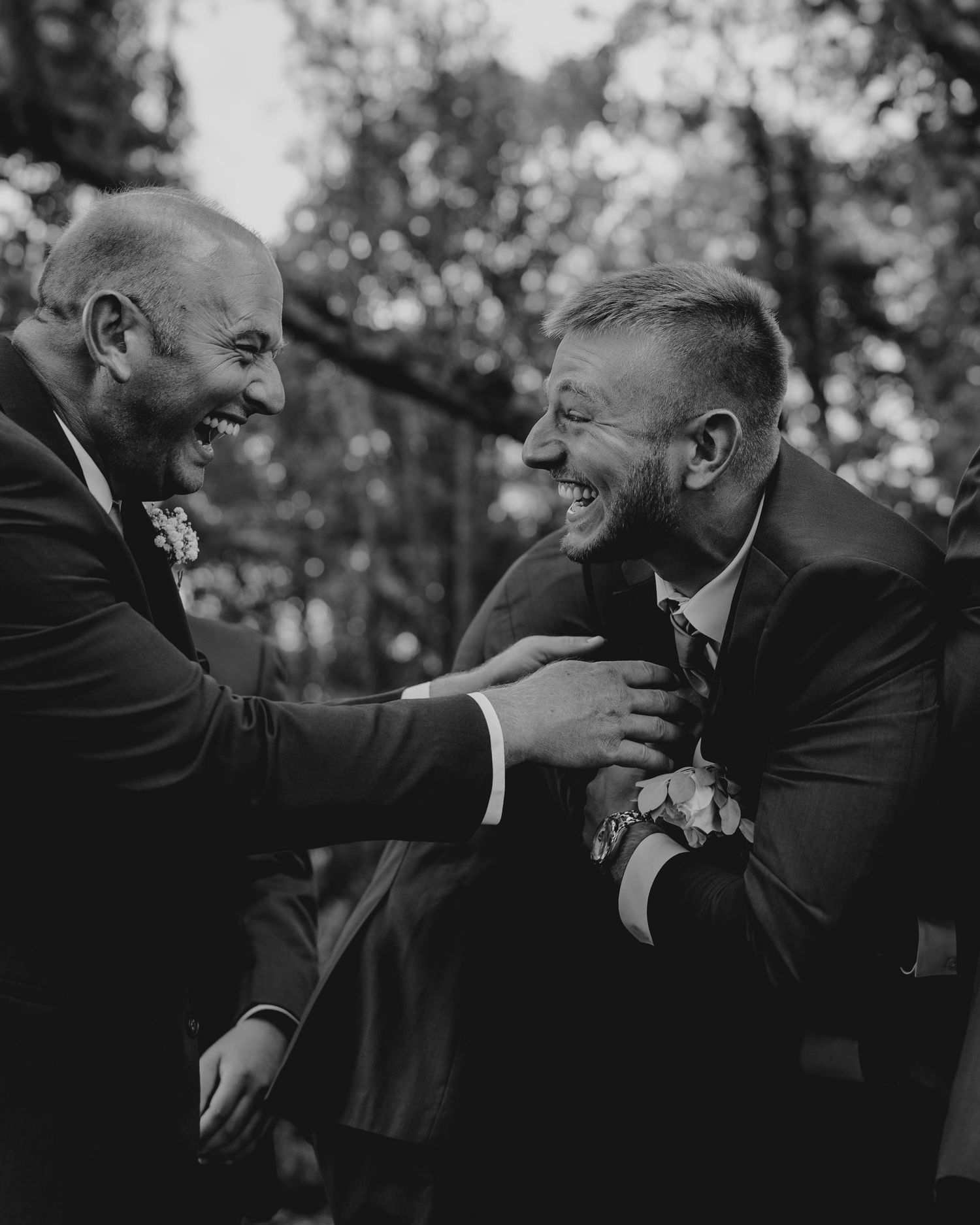 Black and white photo of groom and groomsman laughing joyfully together at outdoor wedding celebration.