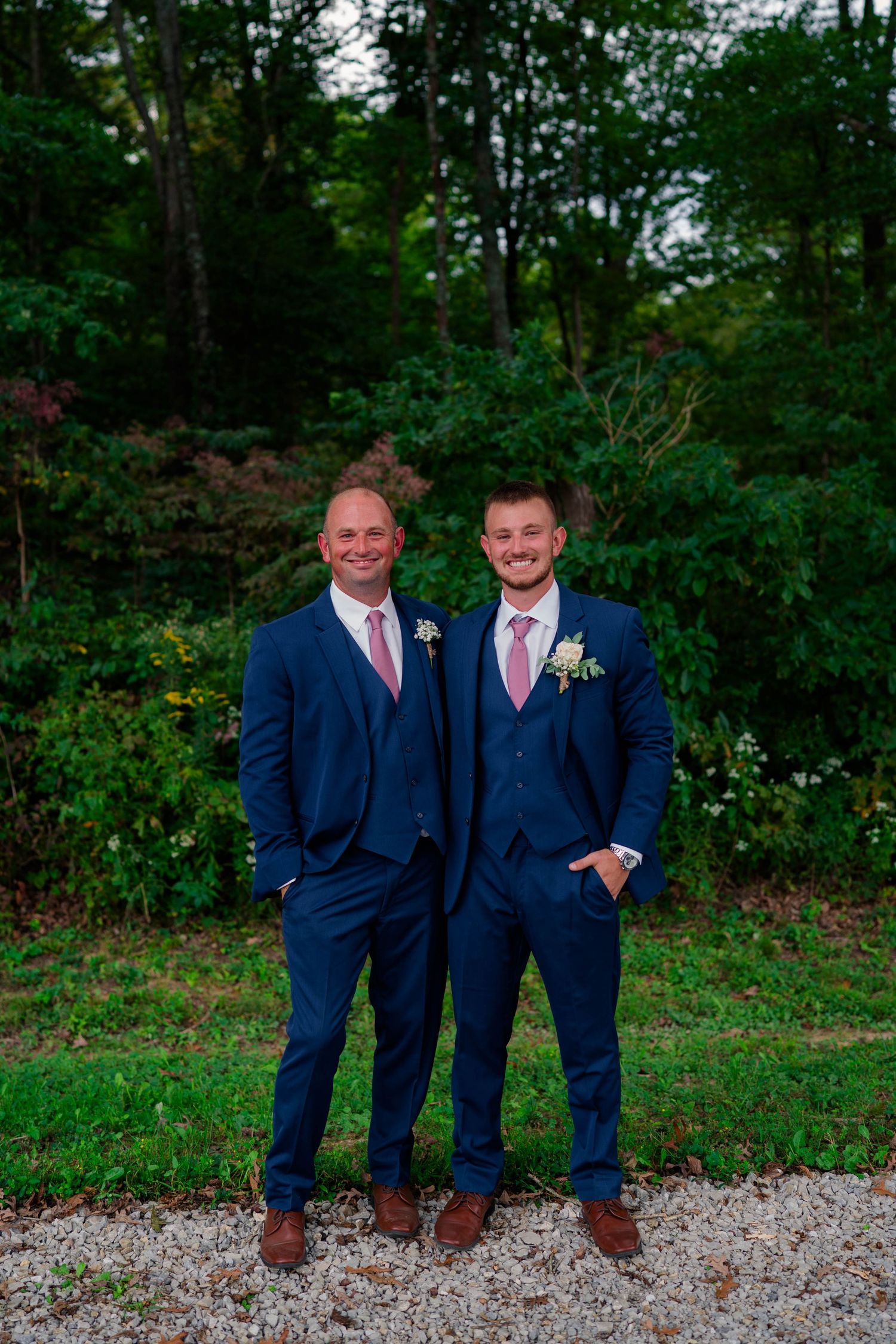 Two groomsmen in navy blue suits with pink ties and boutonnieres outdoors.