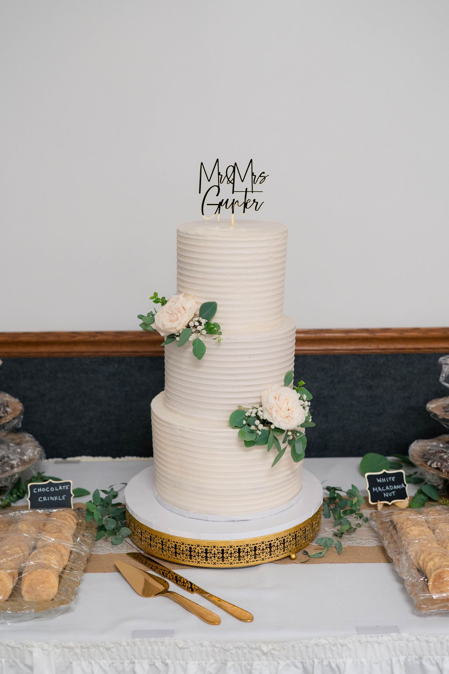 Elegant three-tier white ribbed wedding cake topped with Mr. and Mrs. Gurr topper adorned with white roses and greenery.