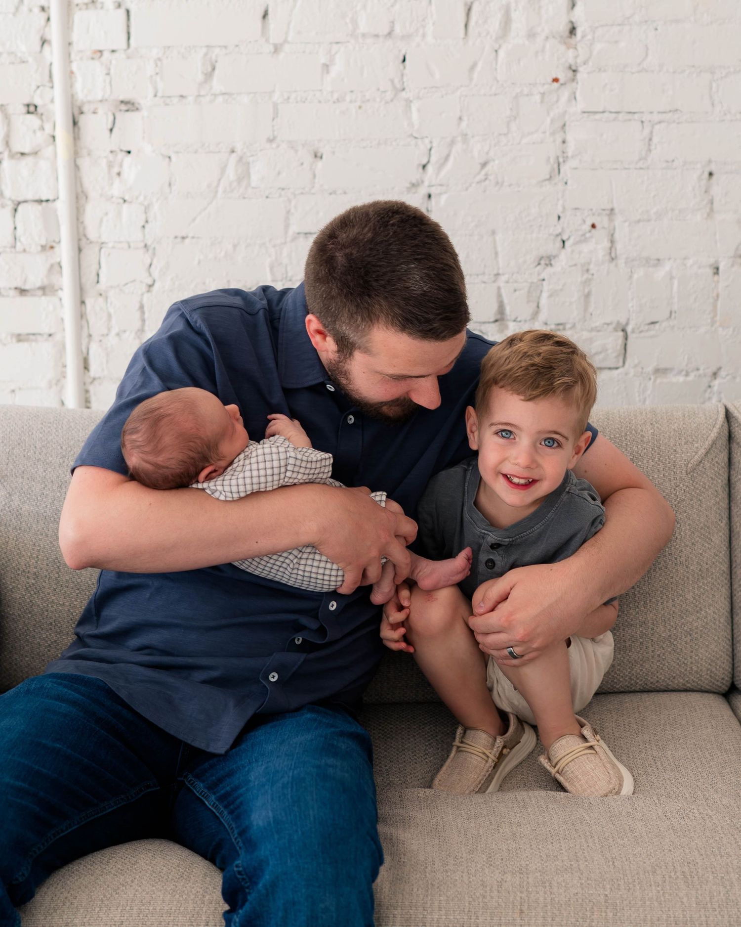 Father holding newborn baby on couch with smiling toddler boy against white brick wall.