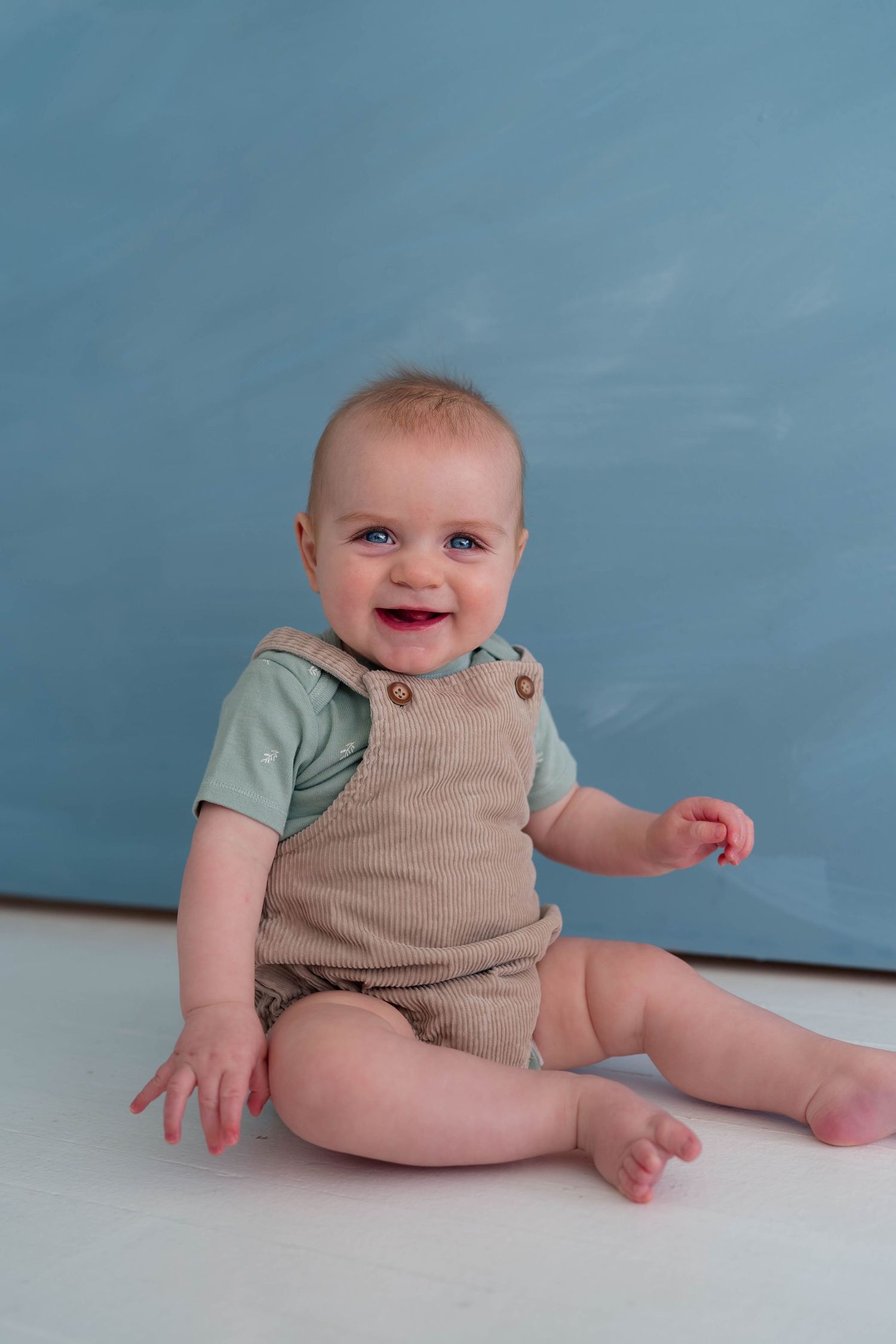 Happy baby boy sitting on white floor in tan overalls and mint shirt with blue painted backdrop.