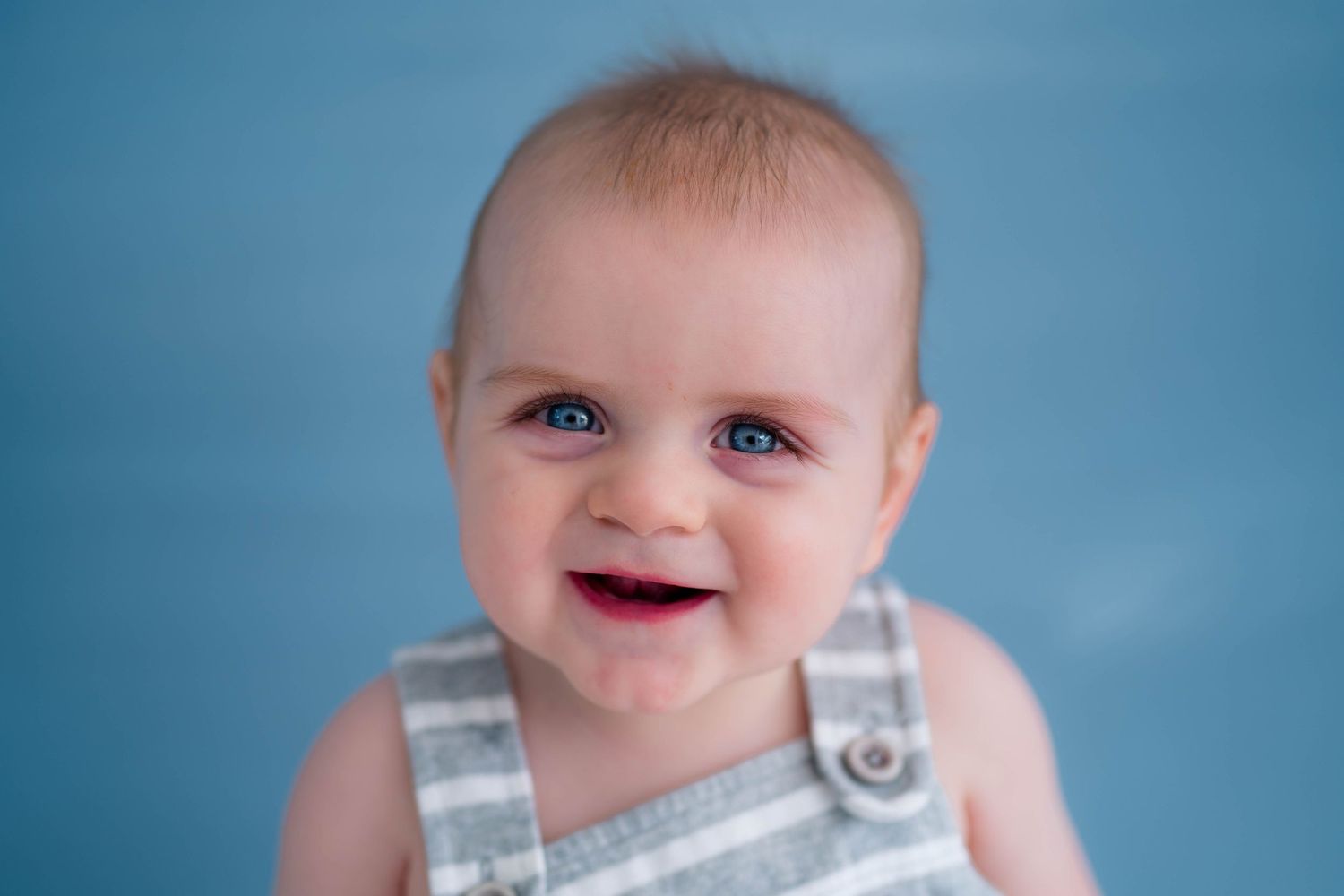 Happy baby with bright blue eyes smiling, wearing gray striped overalls against blue background.