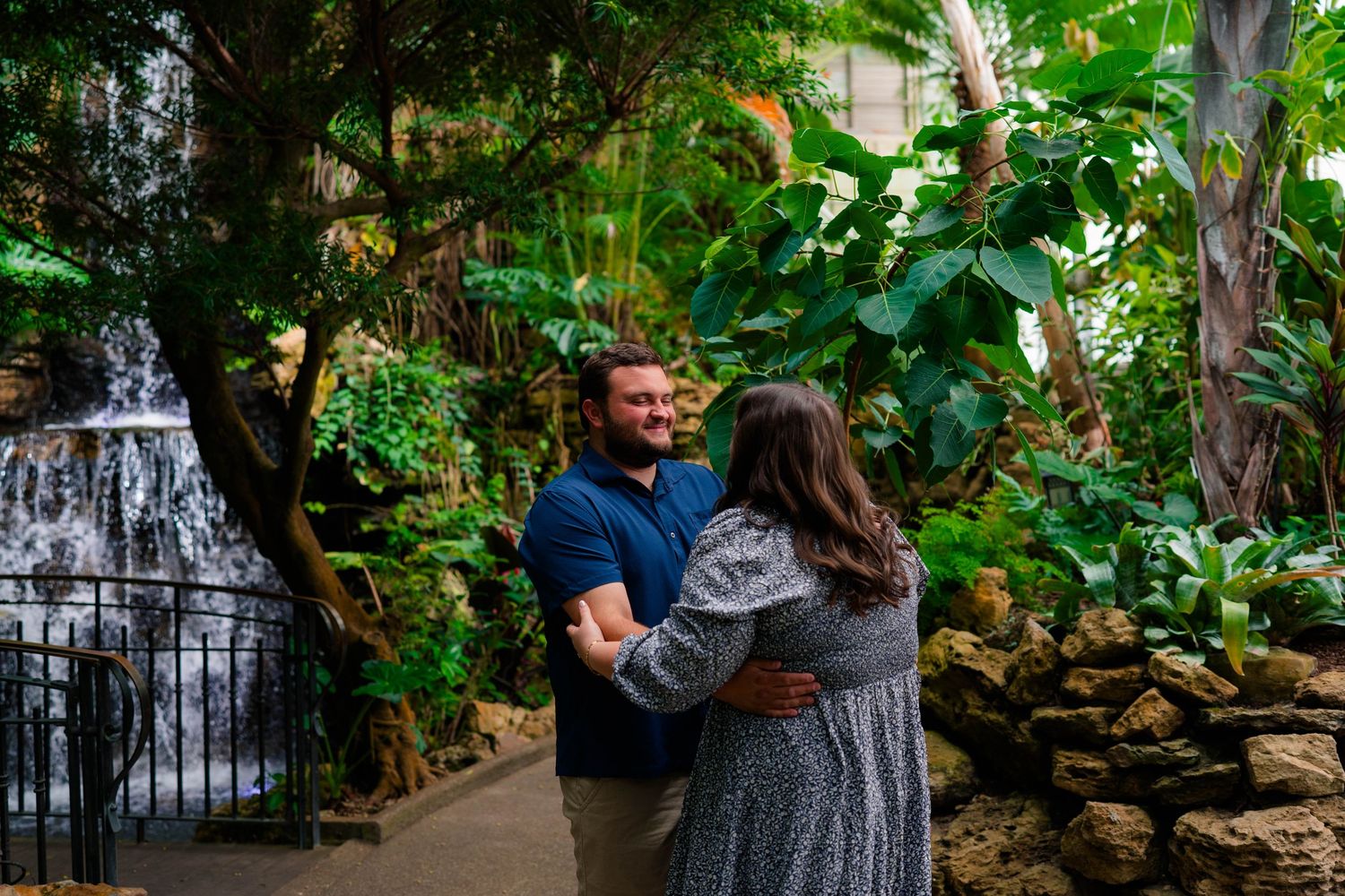 Newly engaged couple smiles and holds hands near a scenic waterfall in a vibrant tropical garden.