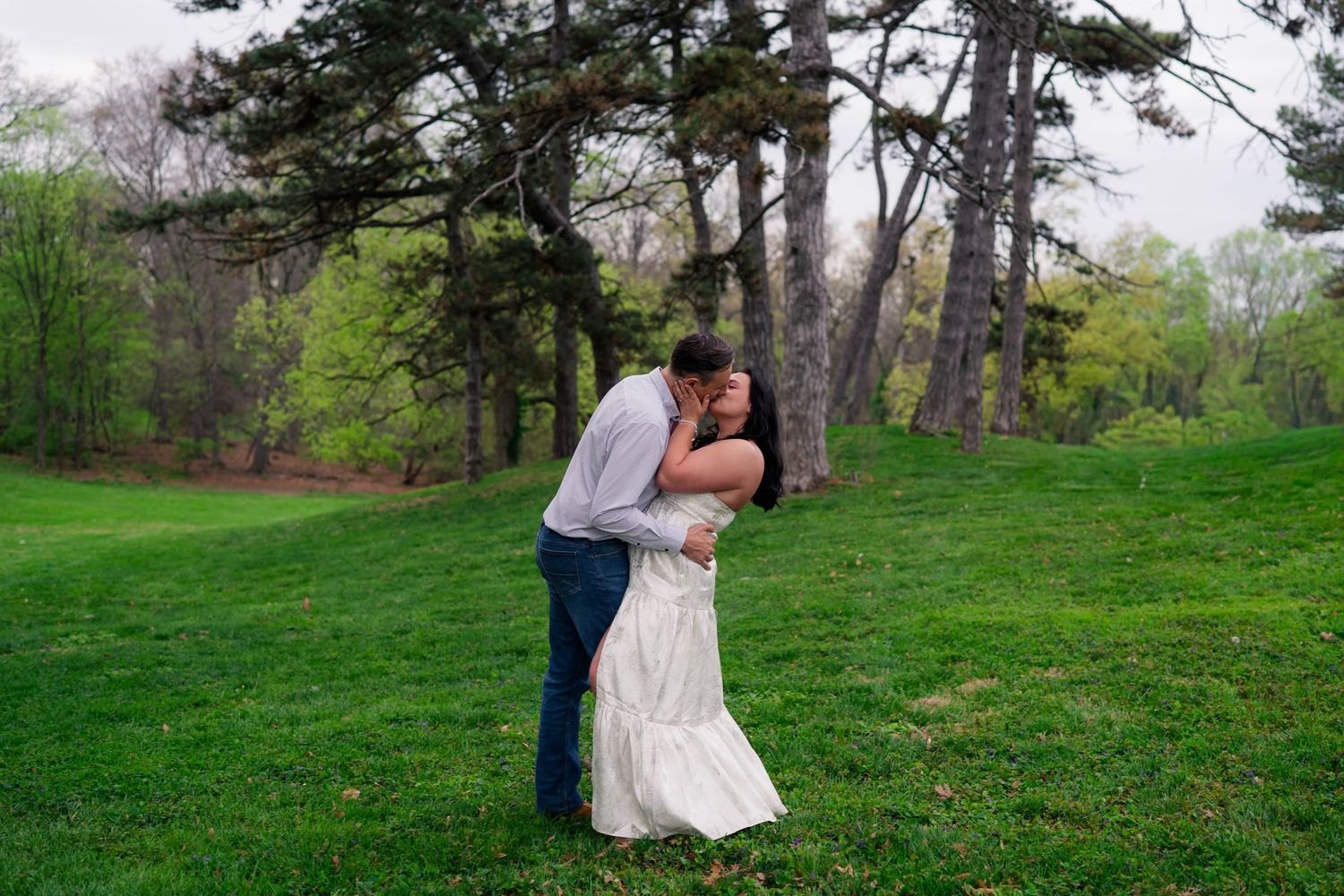 Couple sharing a romantic dip kiss on lush green grass surrounded by tall trees during an outdoor engagement session.