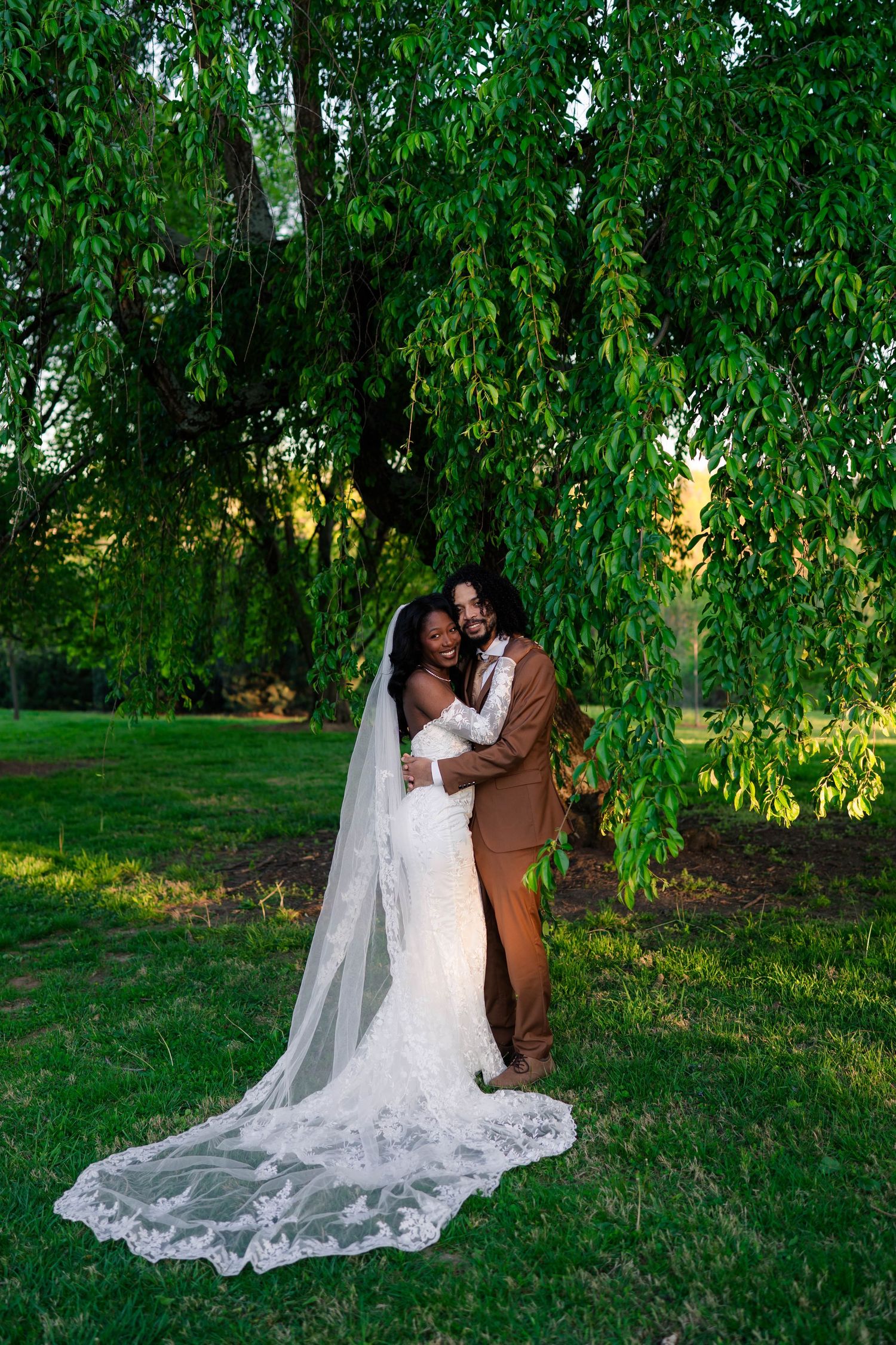 Bride and groom embracing under a weeping willow tree, bride in lace gown with long veil, groom in brown suit.