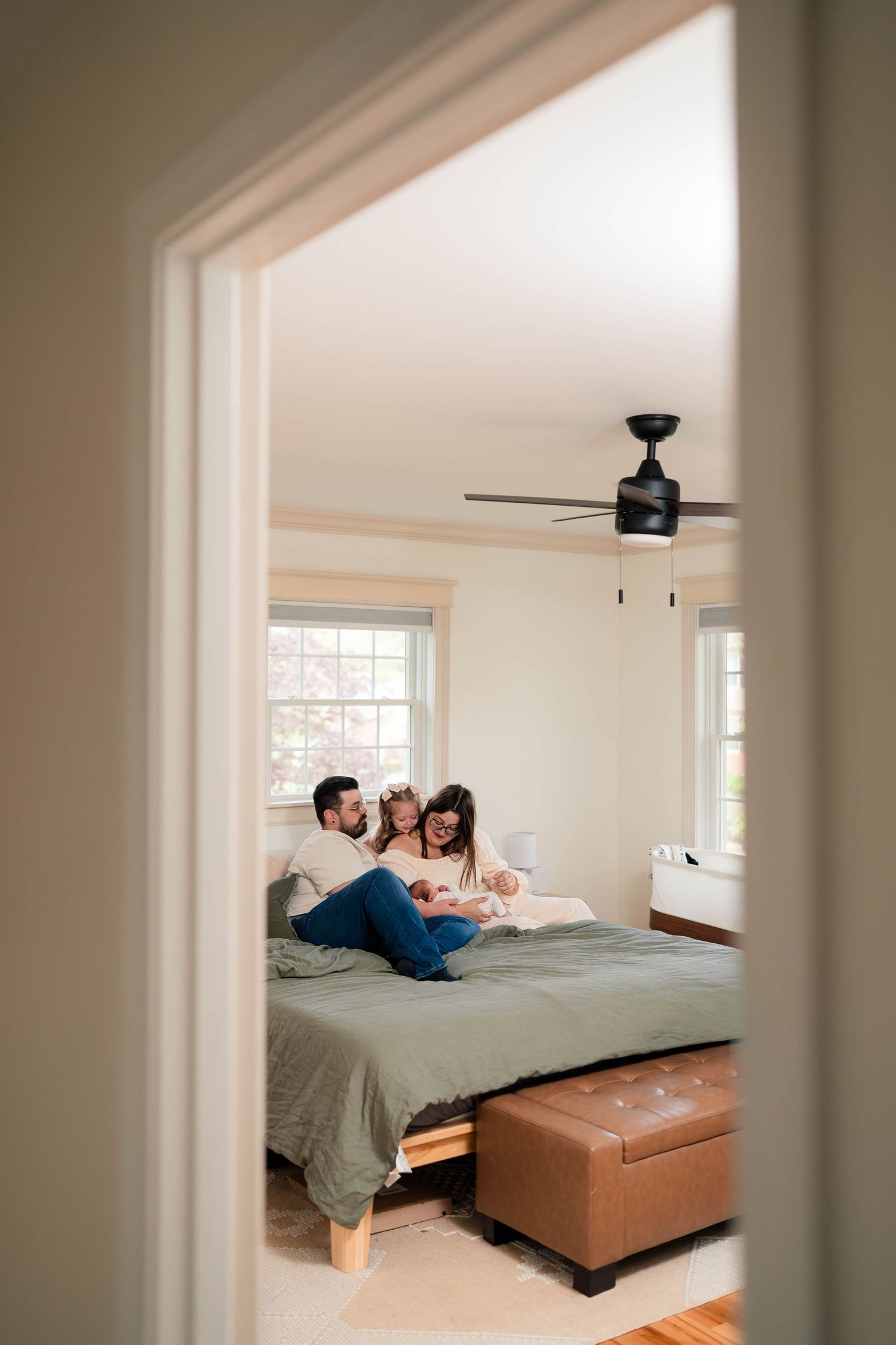 Family of three relaxing together on a green bed in a bright bedroom, viewed through a doorway.