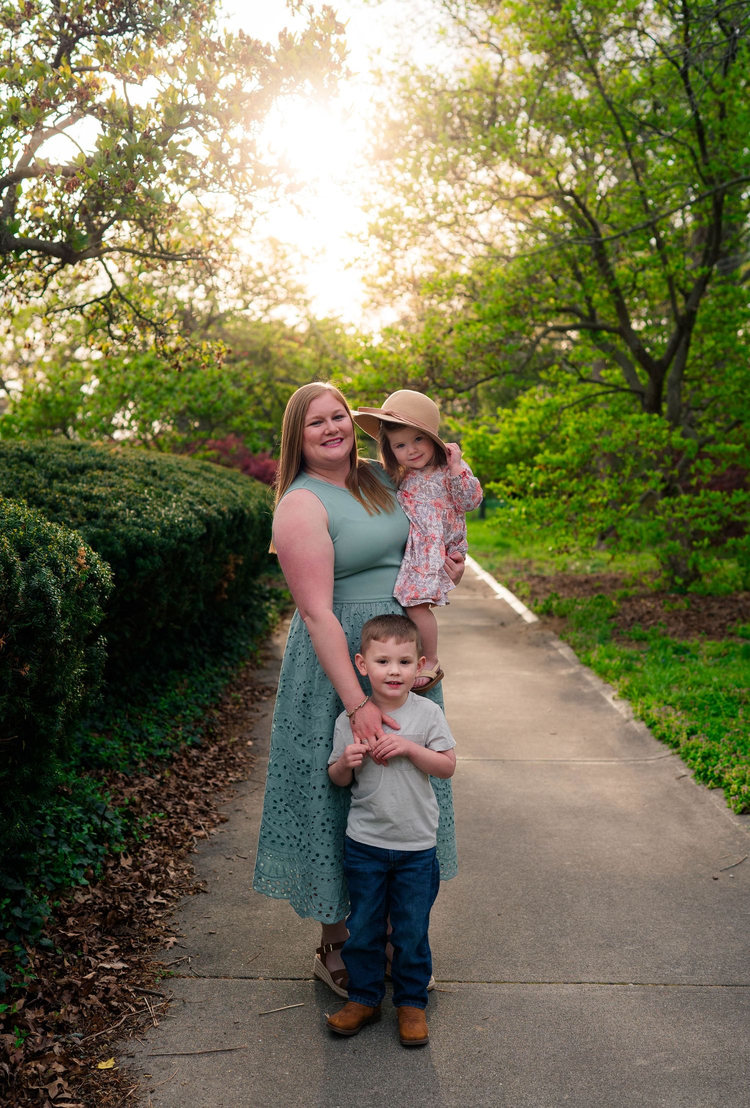 A mother in a teal dress poses with her two young children on a garden path surrounded by lush green trees.
