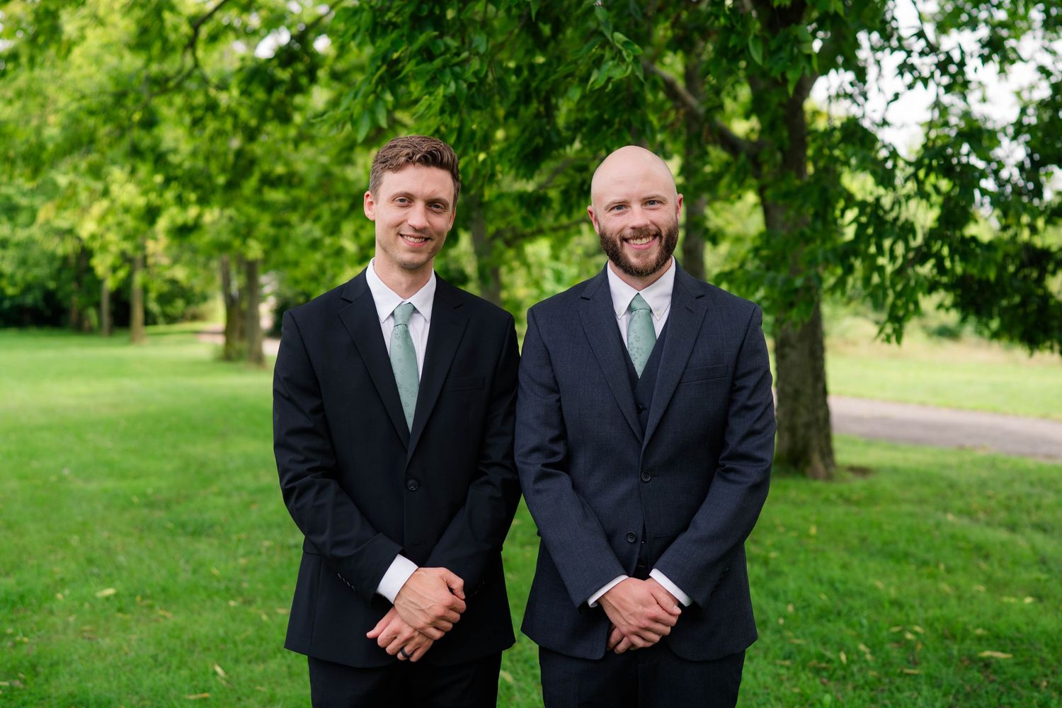 Two men in dark suits with sage green ties smile together on a green lawn at a wedding.