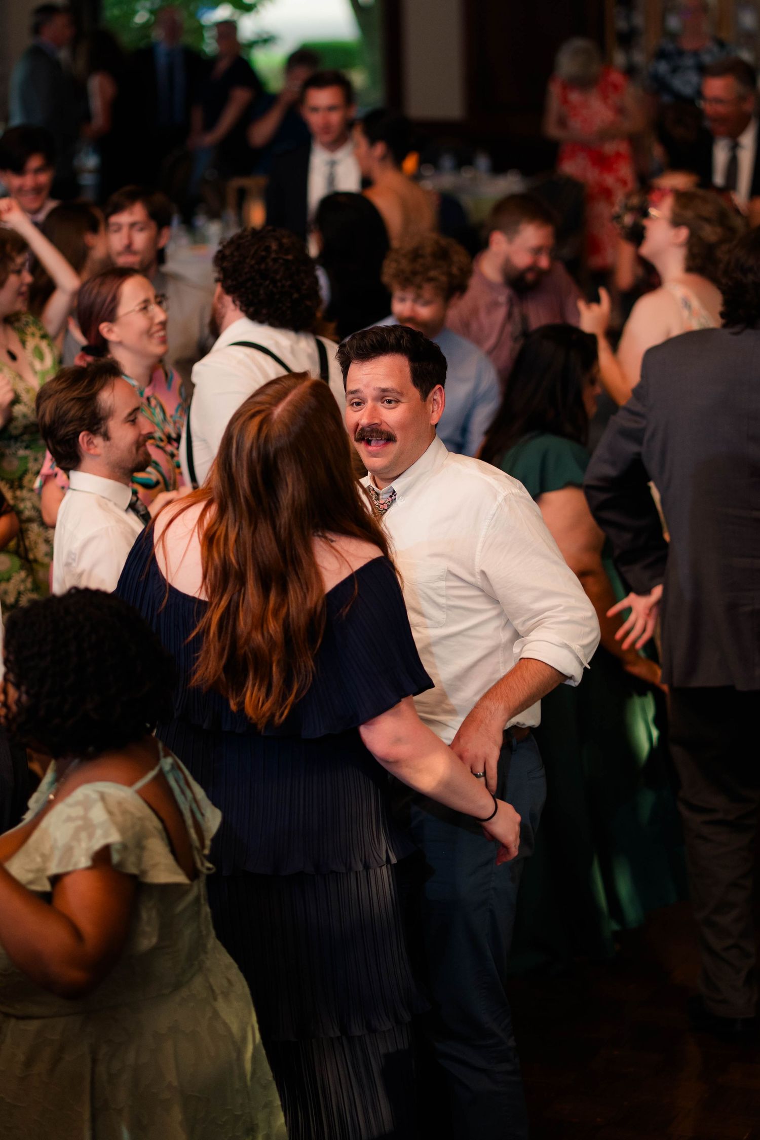 Guests dance together on crowded wedding reception dance floor in warmly lit venue.