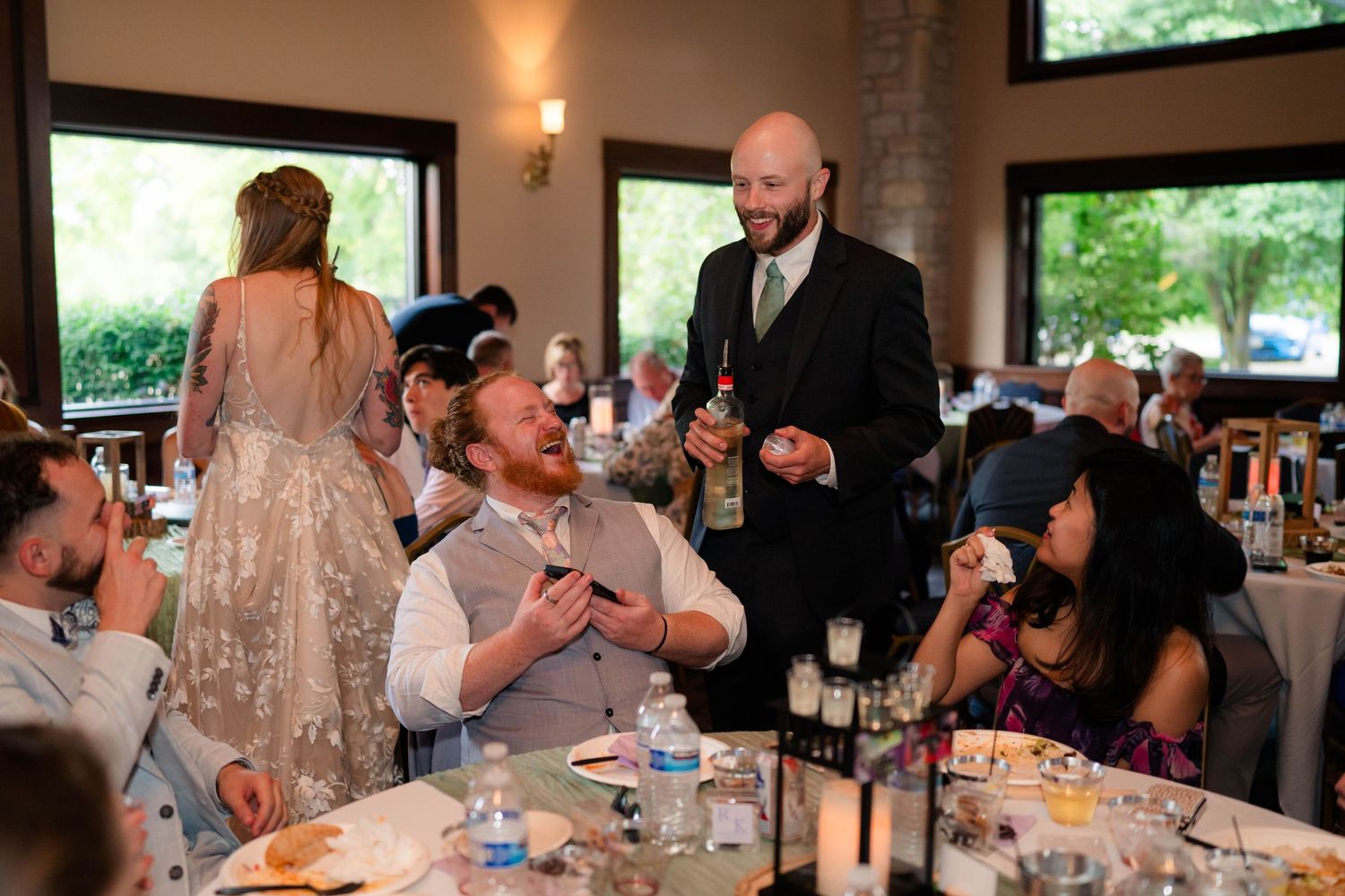 Bald groom mingles with seated wedding reception guests while bride in embellished gown stands in background.