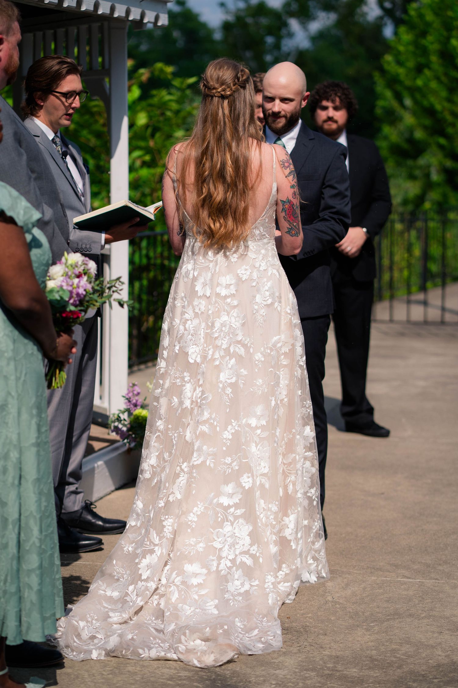Bride in floral lace gown reads vows at outdoor gazebo wedding ceremony with groom and officiant.
