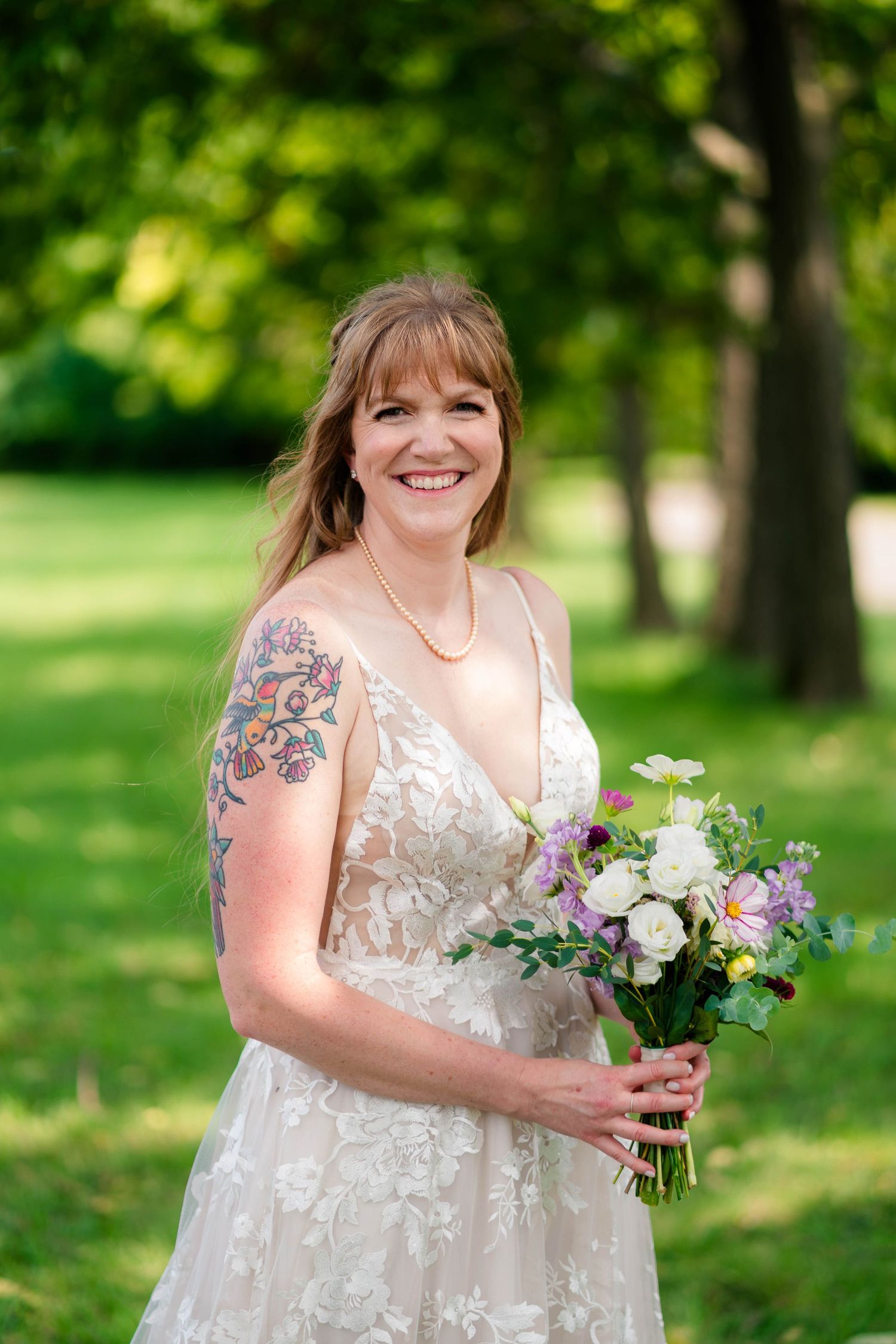Smiling bride in lace gown holding purple and white bouquet with floral arm tattoo in green park.