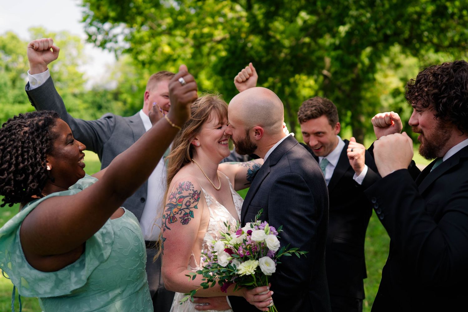 Wedding party cheering as bride and groom share romantic moment surrounded by joyful guests outdoors.